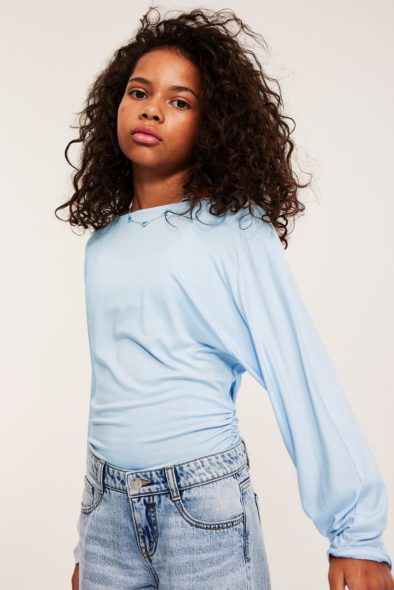 Frontbilde av et barn i lys blå langermet topp, lyse jeans og sølvhalskjede.