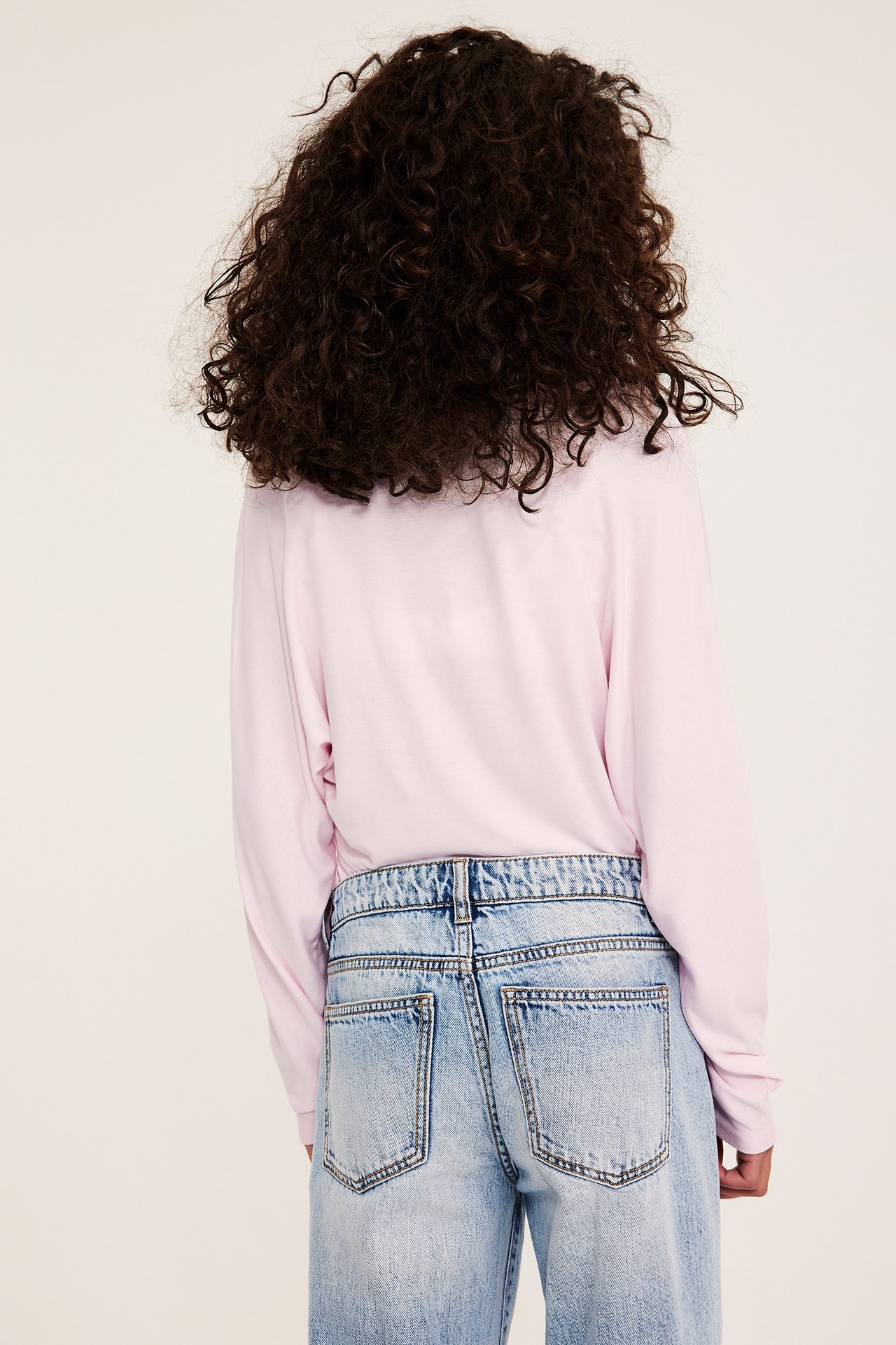 Bakfra, et barn i lyserosa ensfarget langermet topp og lyseblå slitte denimjeans.
