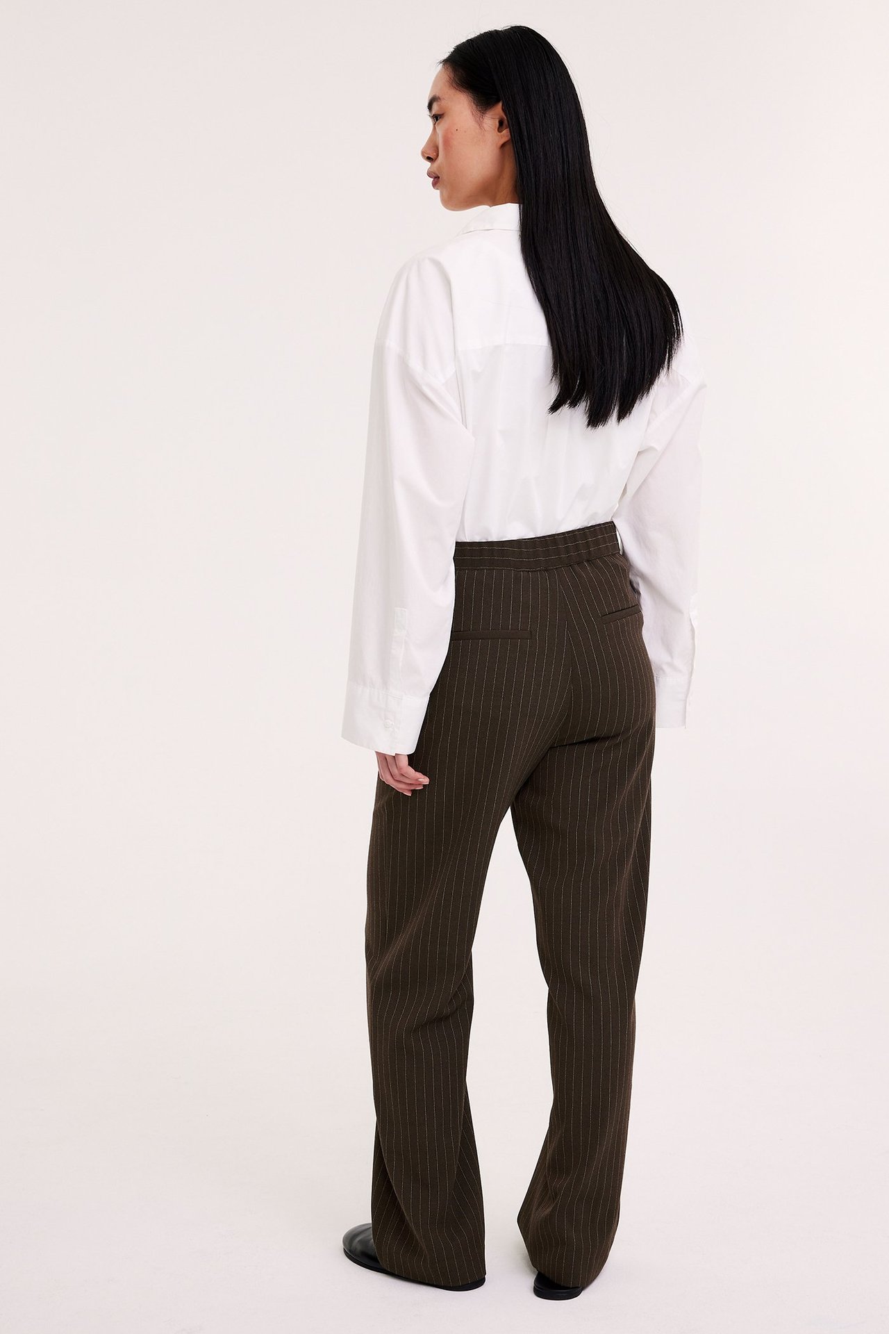 Bakifrån syns en kvinna i mörkbruna randiga slouchy kostymbyxor och en vit skjorta.