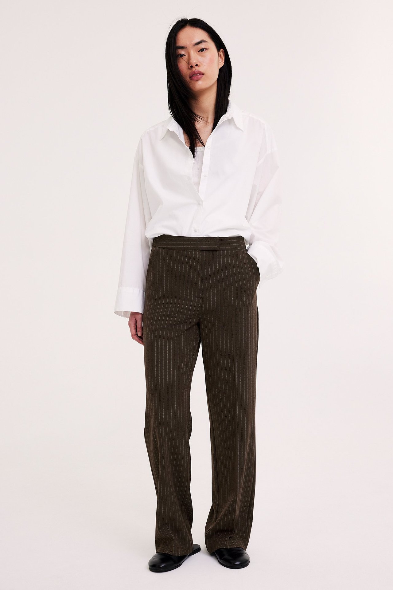Forfra: kvinne i mørkebrune stripete dressbukser med vide ben, hvit oversized skjorte og svarte flate sko.
