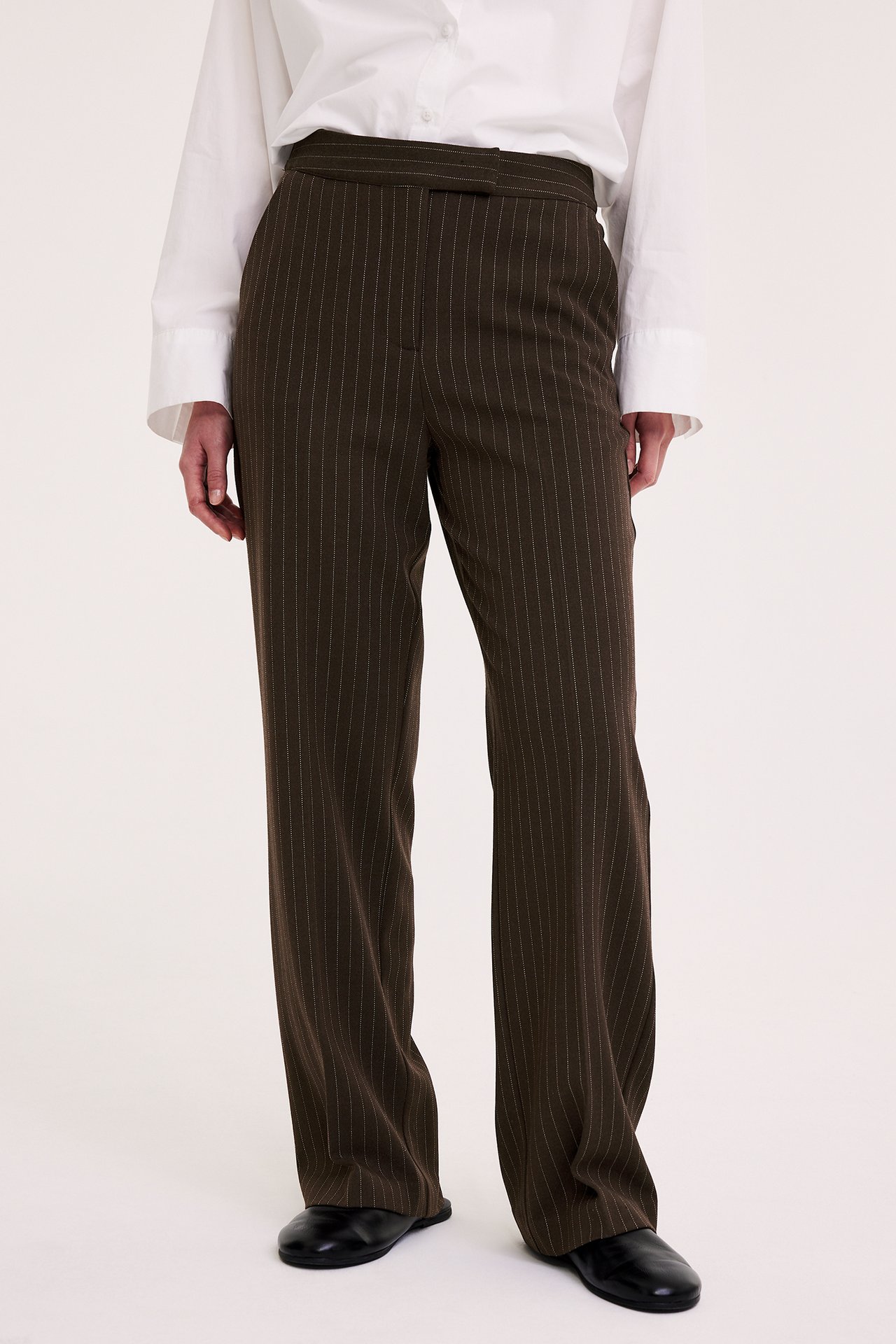 Frontvy av en kvinna i mörkbruna randiga slouchy kostymbyxor, stylade med vit skjorta och svarta platta skor.