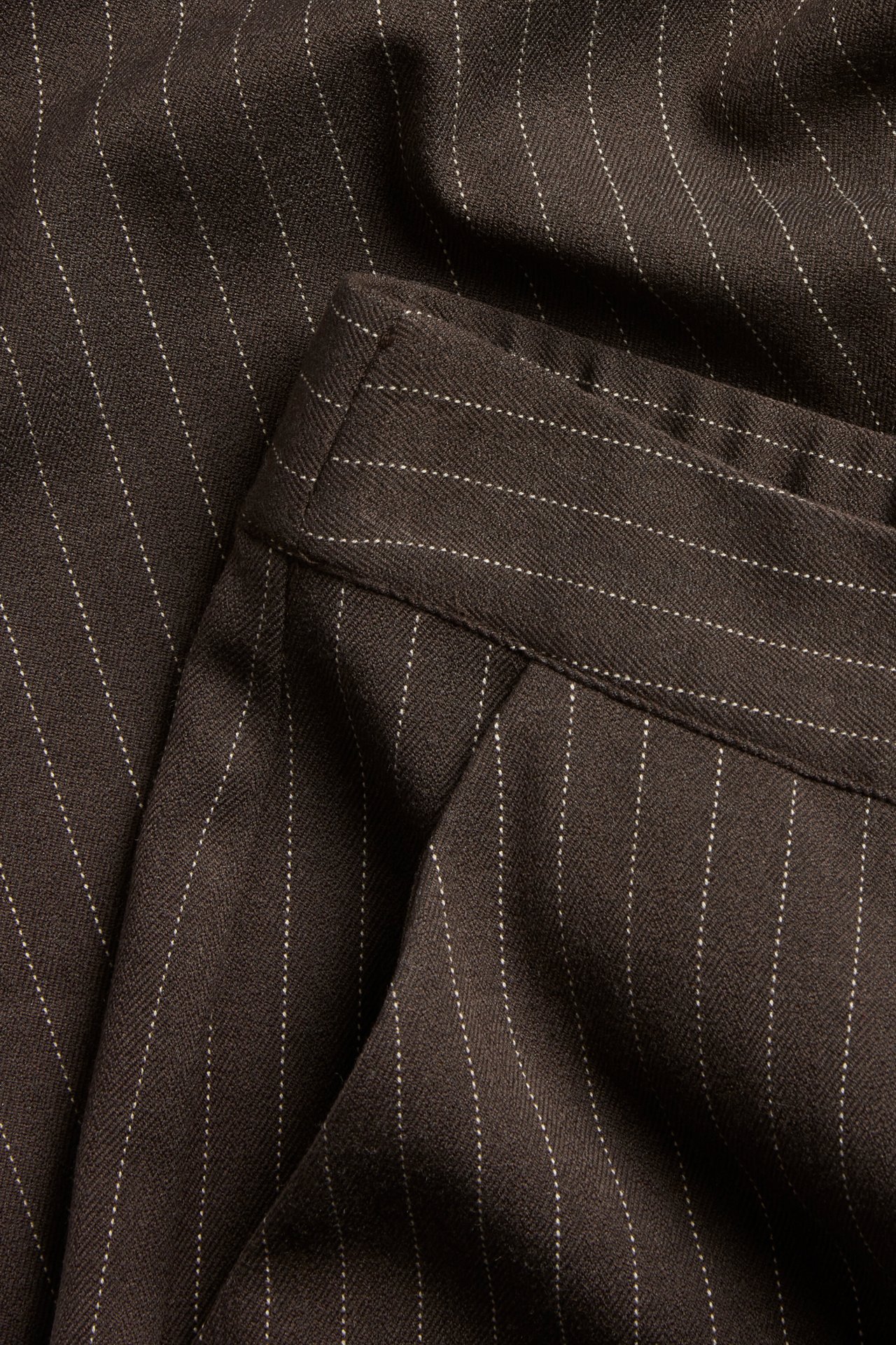 Zbliżenie na ciemnobrązowe, prążkowane spodnie garniturowe o fasonie slouchy z widocznym pasem.