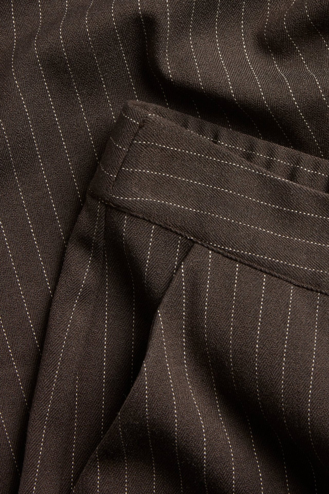 Zbliżenie na ciemnobrązowe, prążkowane spodnie garniturowe o fasonie slouchy z widocznym pasem.