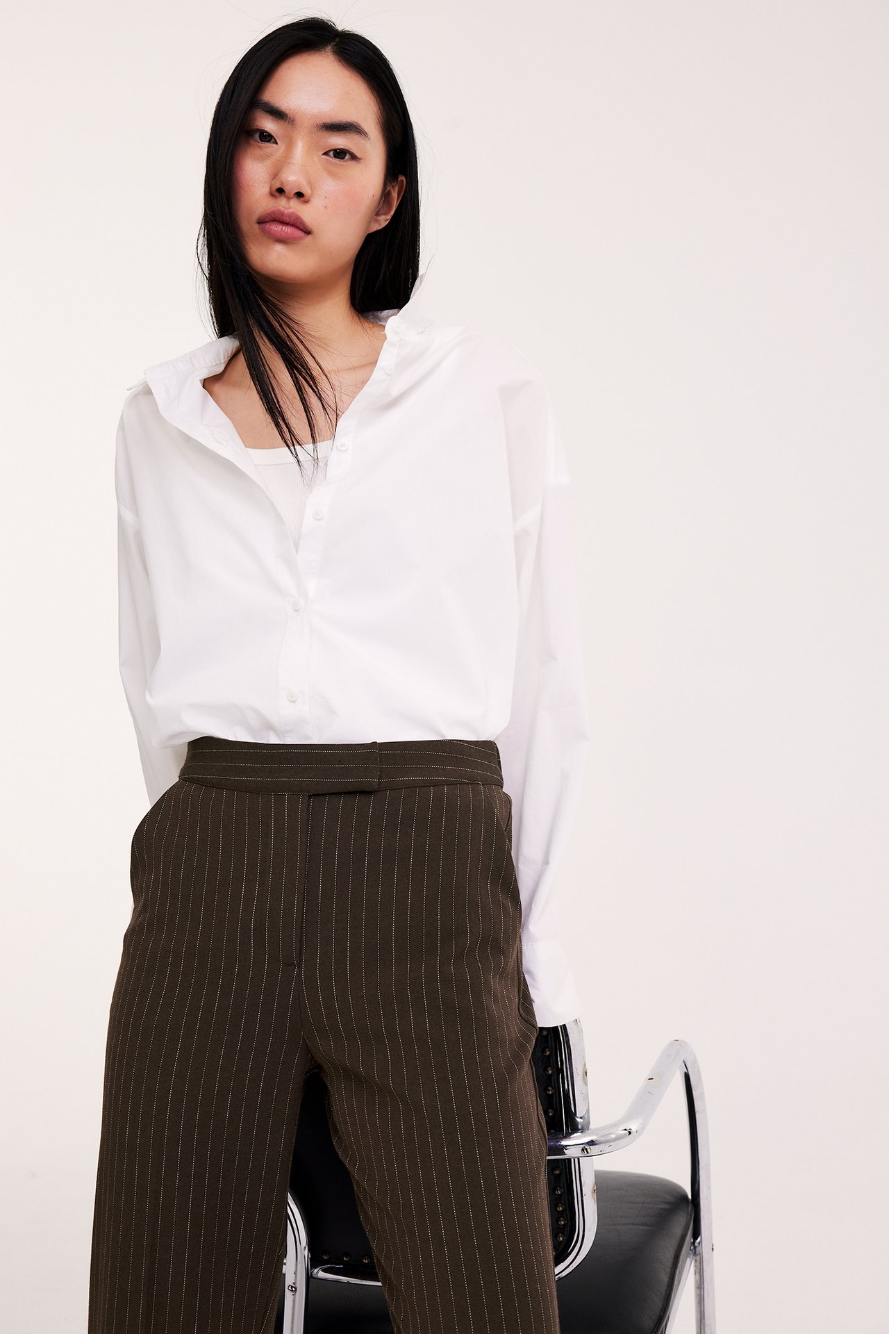 Framifrån, kvinna i mörkbruna randiga slouchy kostymbyxor och en vit skjorta över ett vitt linne.