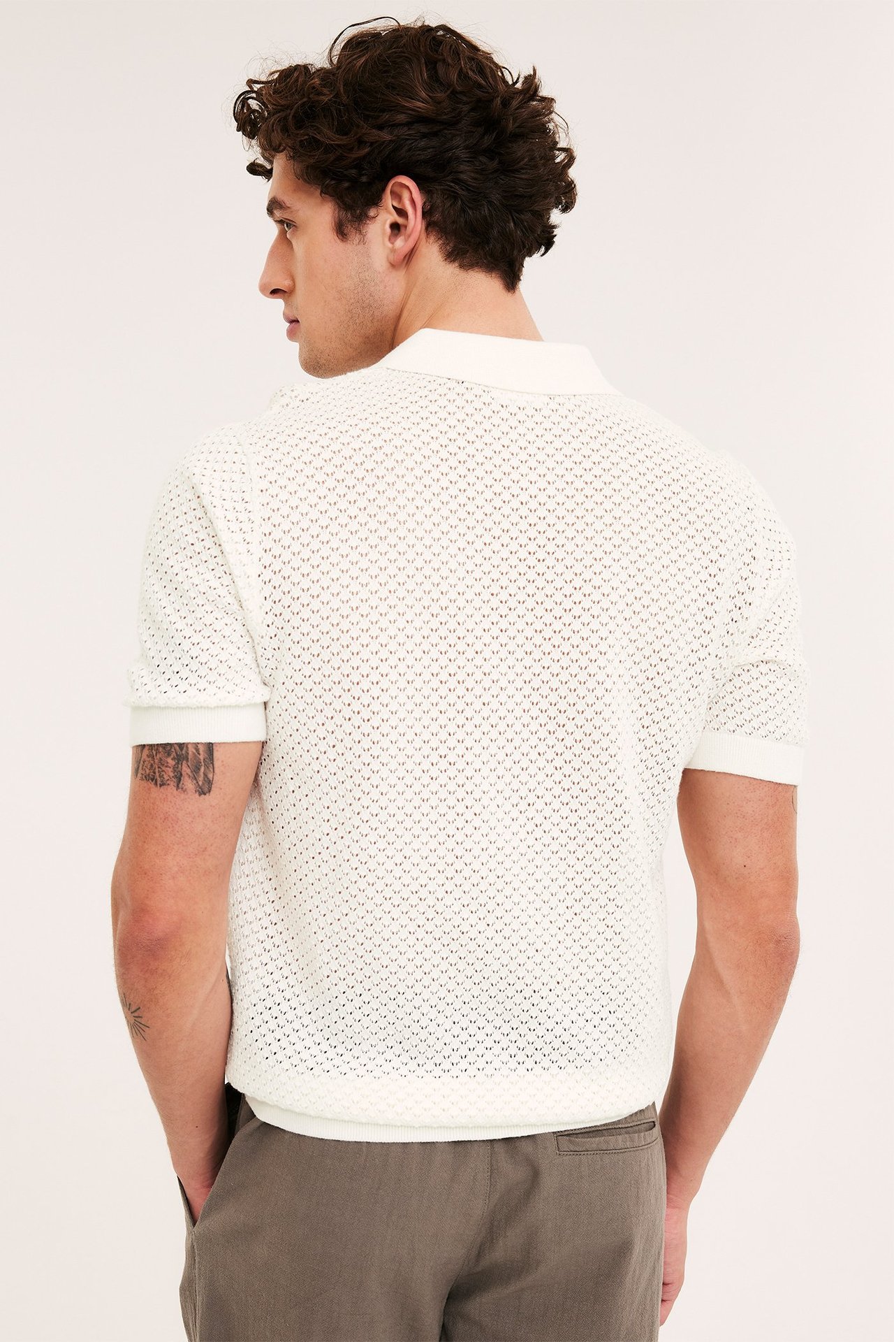 Man bakifrån i en offwhite kortärmad stickad pikétröja med hålmönster och mörkgråa byxor.