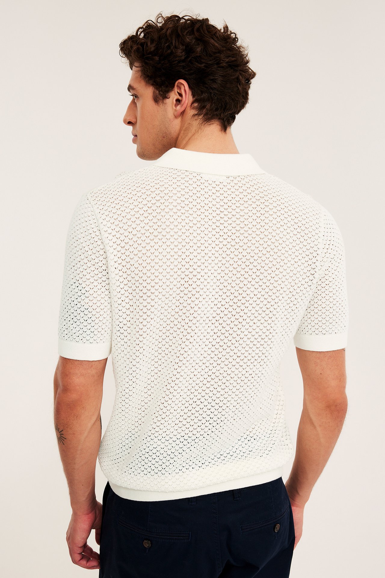 Ryggvy av en man i en råvit hålstickad kortärmad pikétröja och mörkblå byxor.