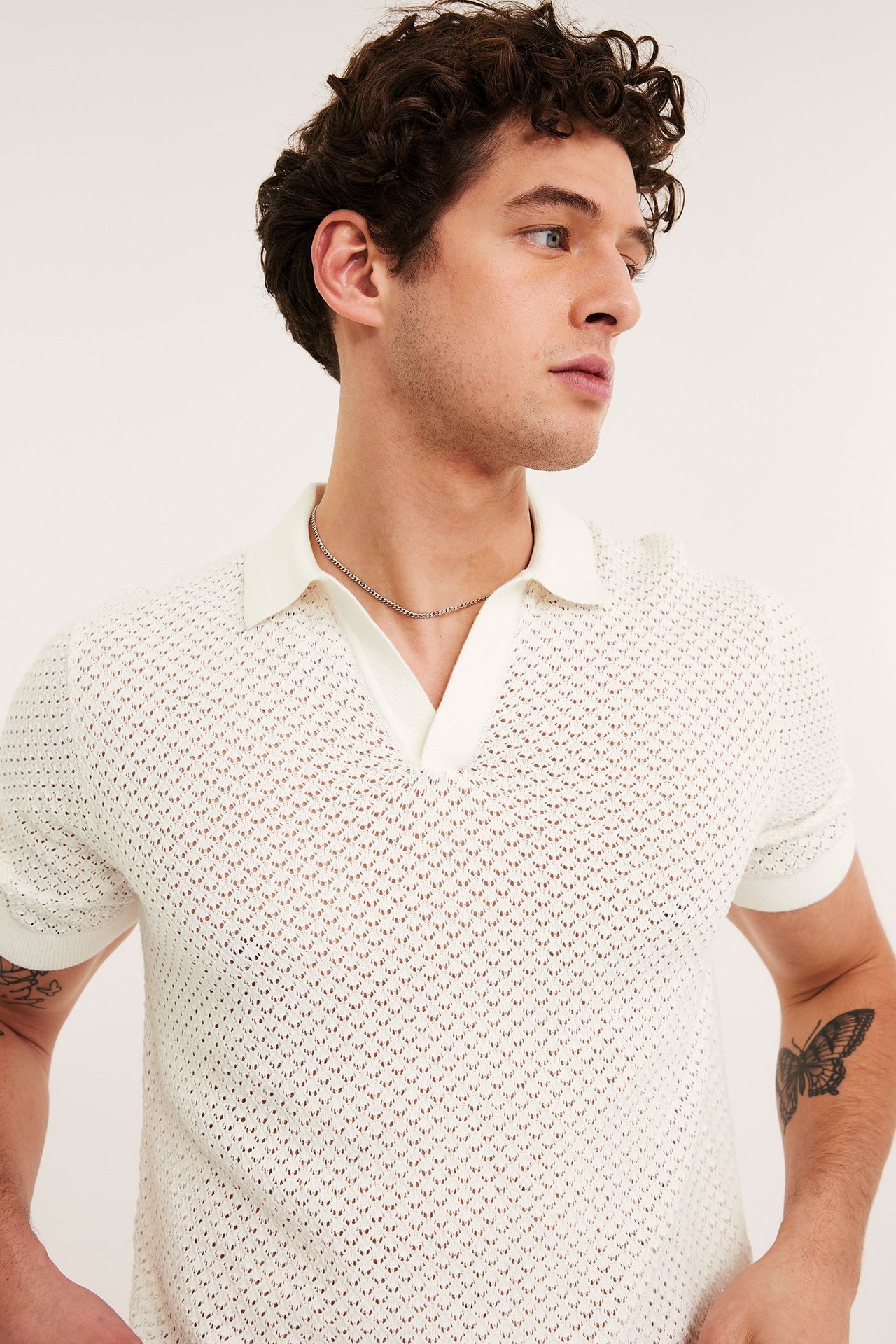 Närbild på en man i en offwhite hålstickad kortärmad pikétröja och ett silverfärgat halsband.