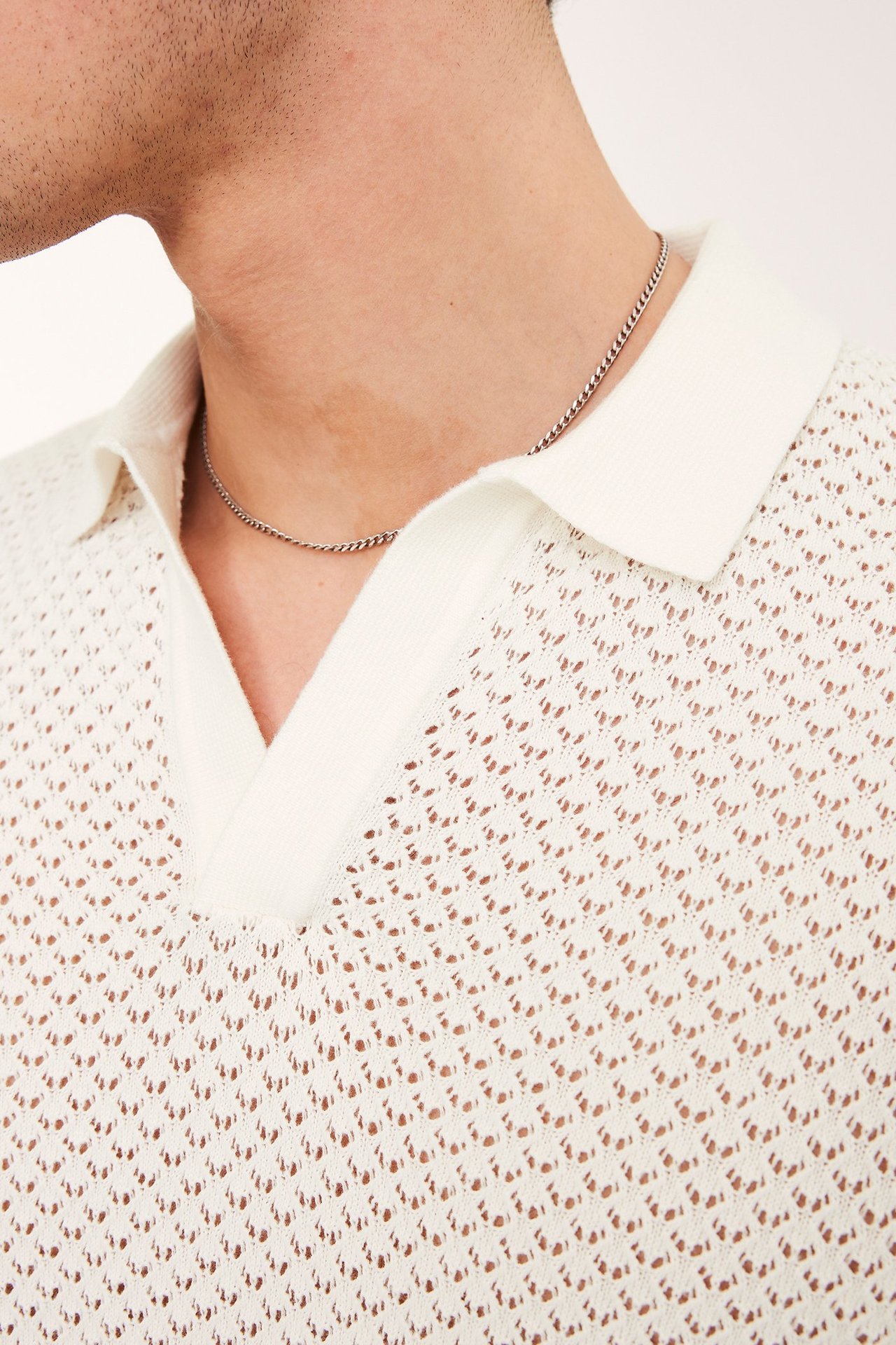 Närbild på en man i en naturvit hålstickad kortärmad pikétröja och ett silverfärgat halsband.