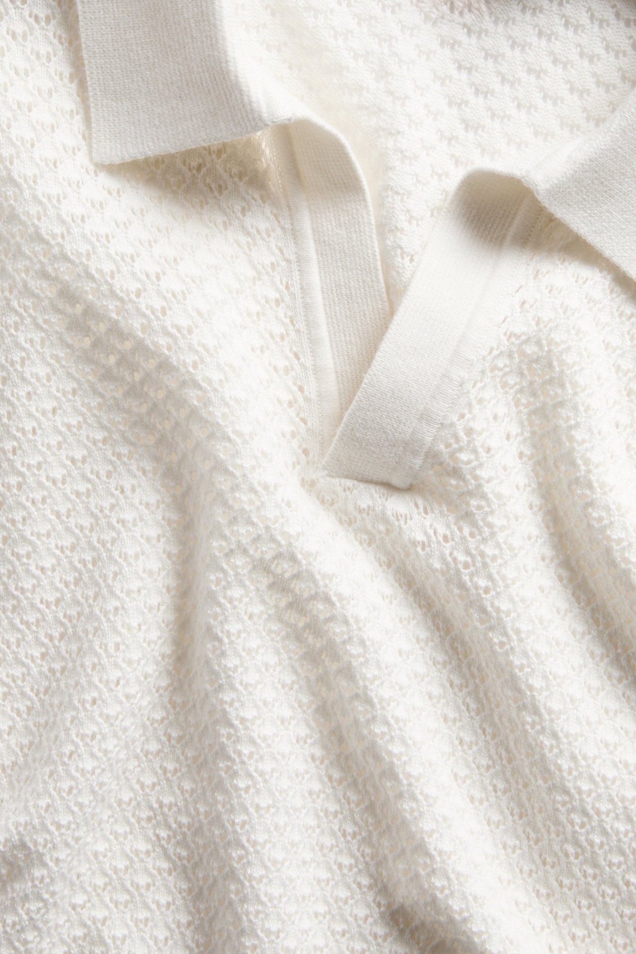 Närbild på en off-white stickad pikétröja i bomull med diskret hålmönster och krage.