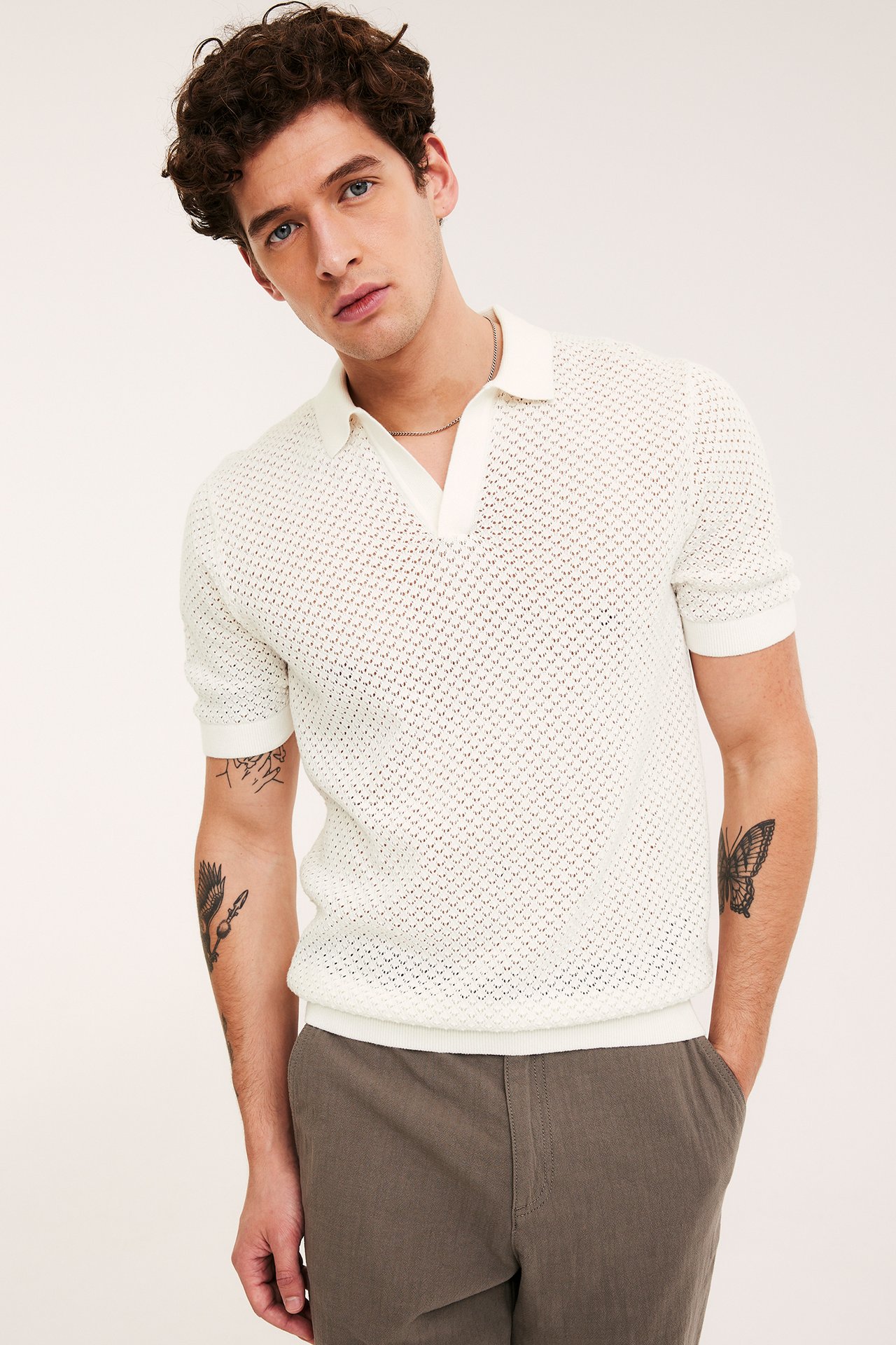 Framifrån, man i offwhite hålstickad kortärmad pikétröja, silverhalsband och mörkgrå byxor.