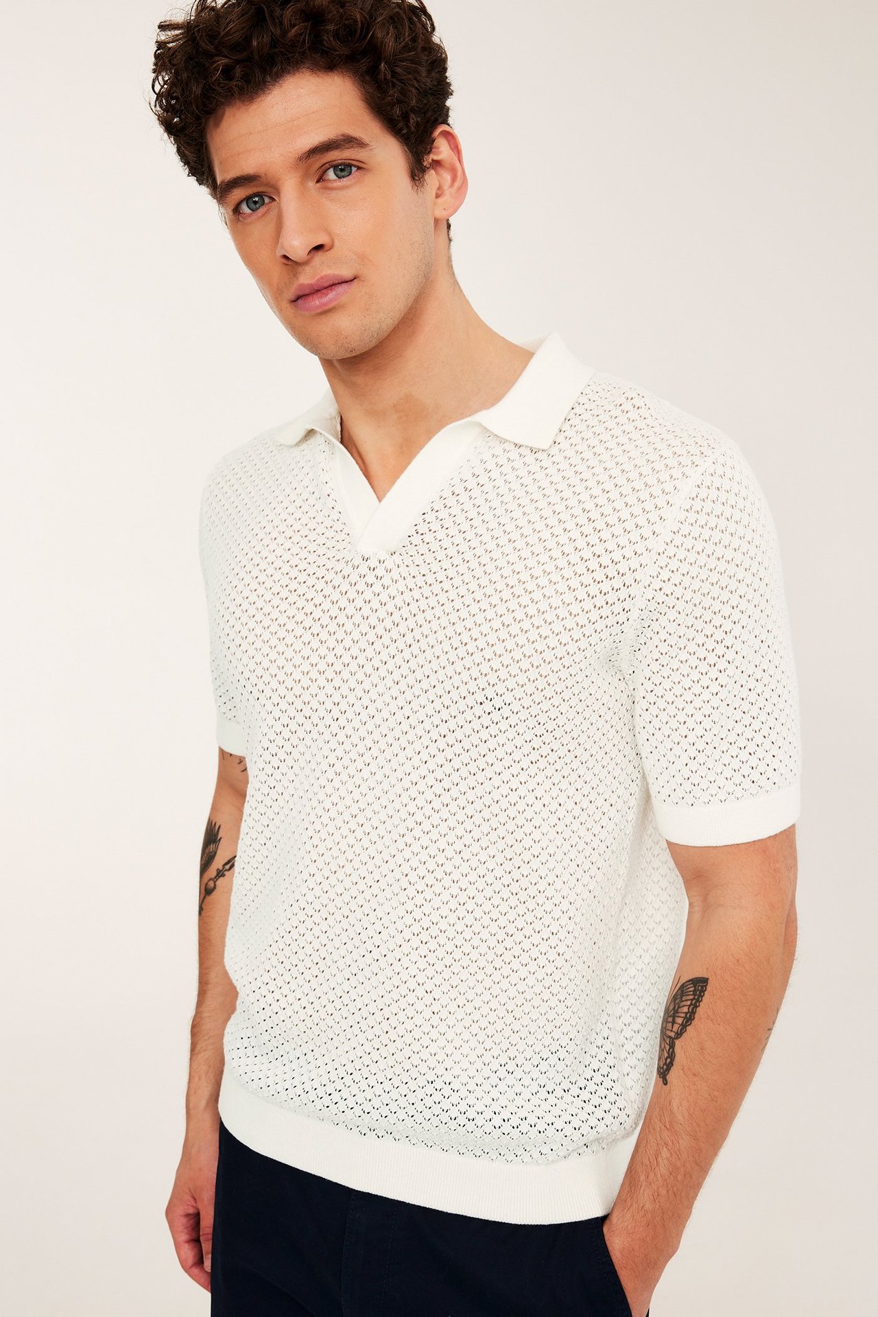 Edestä: mies yllään luonnonvalkoinen neulottu lyhythihainen poolopaita reiäkäs kuviolla ja tummansiniset housut.