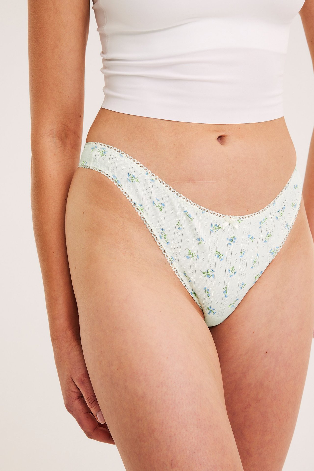 Edestäpäin, nainen yllään vaaleanvihreät kukkakuvioiset pointelle-stringit luonnonvalkoisella pitsireunalla ja pienellä rusetilla.