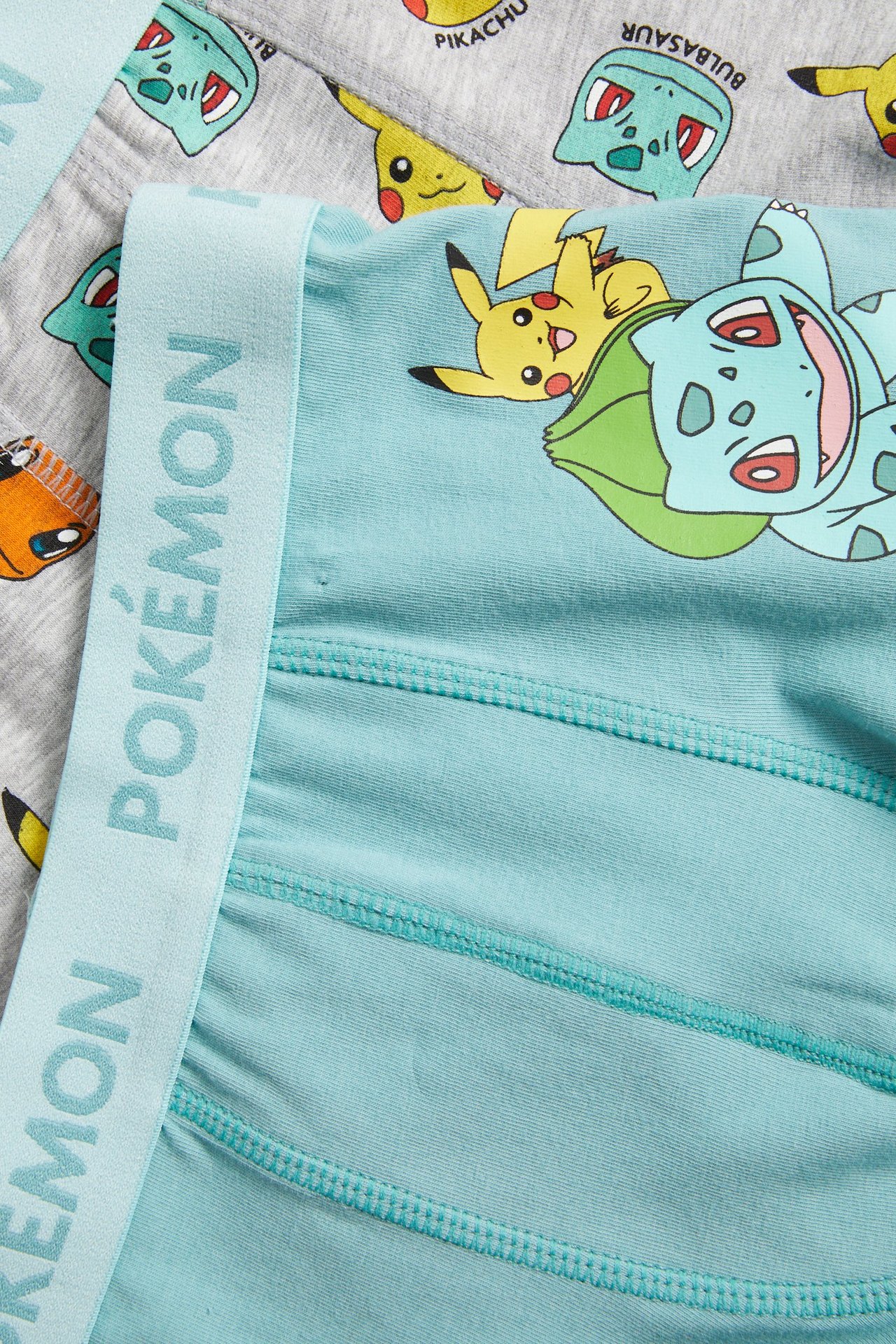Zbliżenie na jasnoniebieskie bokserki dziecięce z Bulbasaur, Pikachu i POKÉMON-paskiem, na szarych bokserkach Pokémon.