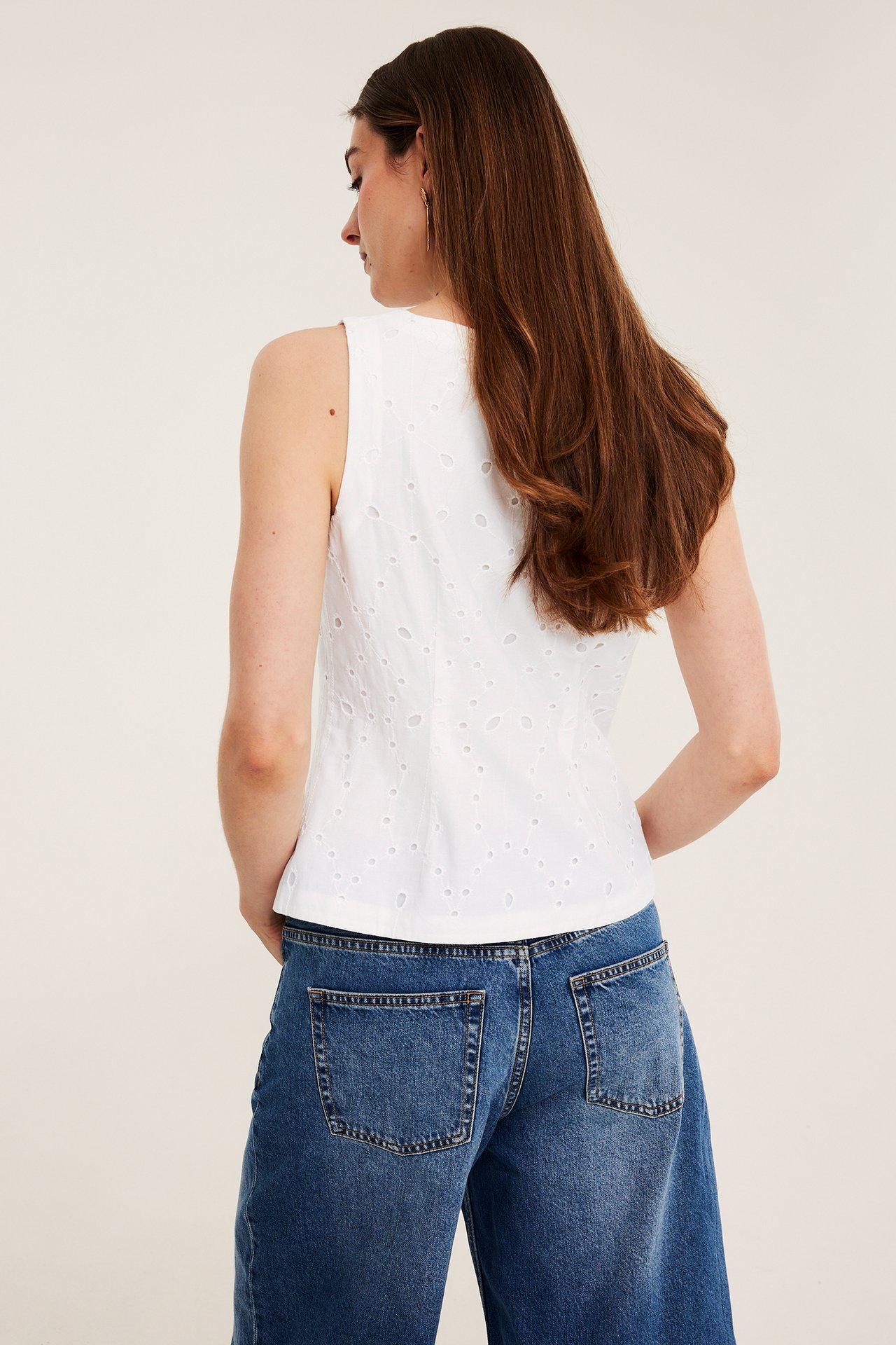 Takaapäin: nainen valkoisessa hihattomassa broderie anglaise -liivissä ja sinisissä leveissä farkuissa.