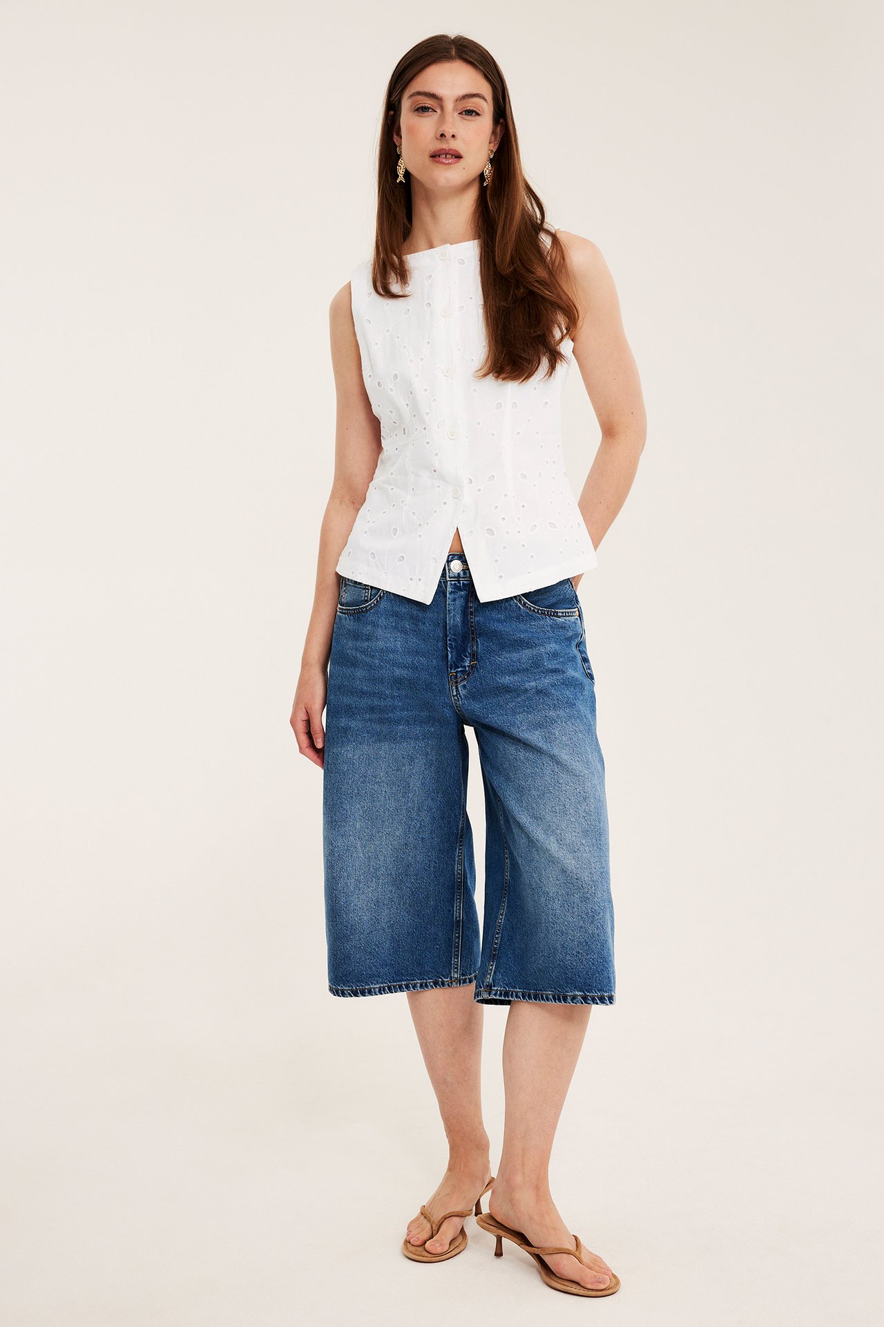 Nainen edestäpäin, valkoinen broderie anglaise -liivi, siniset farkkuculottesit ja ruskeat korkokengät.