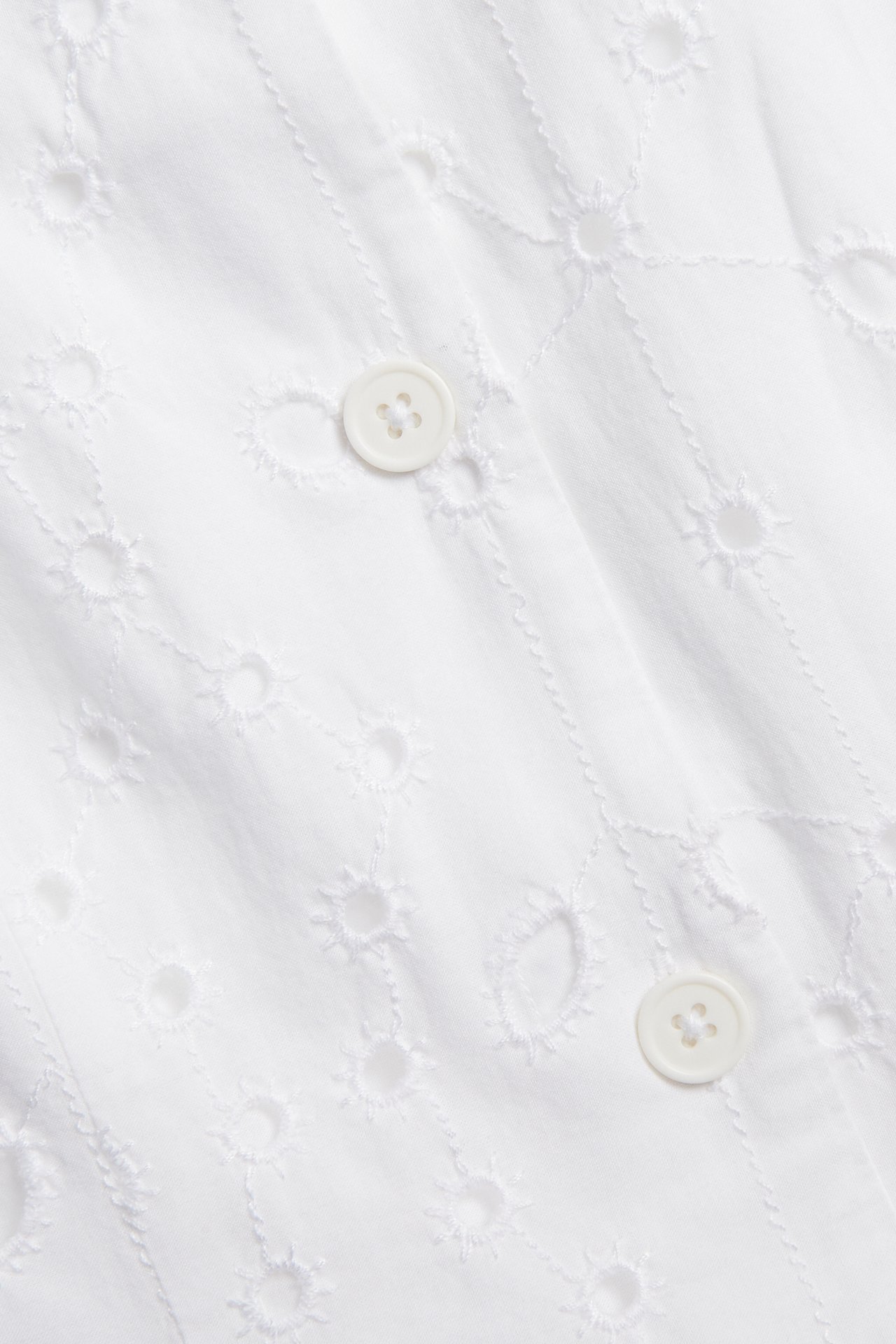 Närbild av en vit väst i broderie anglaise av bomull med knappar fram.