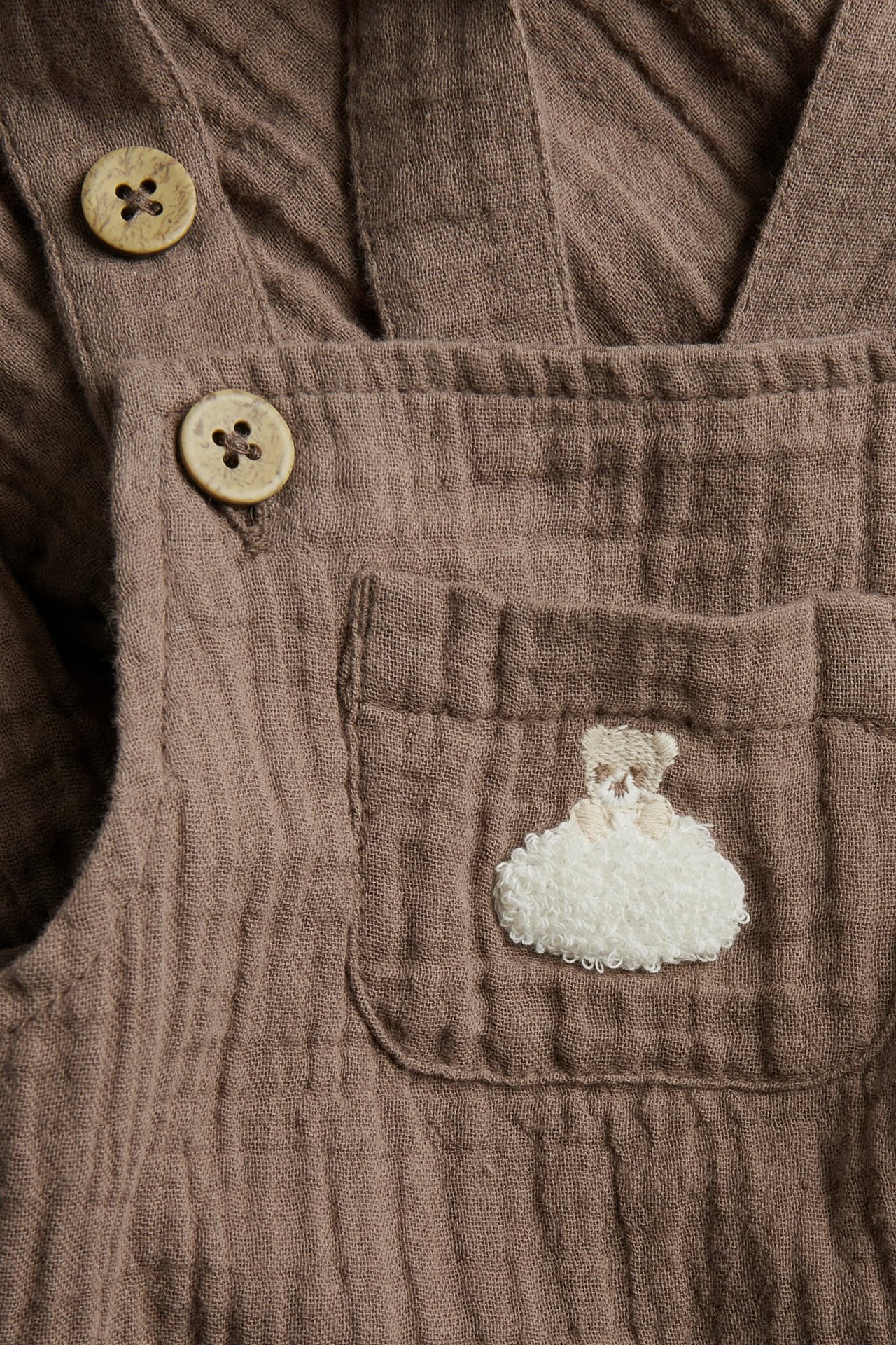 Närbild på bruna hängselbyxor i muslin med träknappar och broderad nalle med moln.