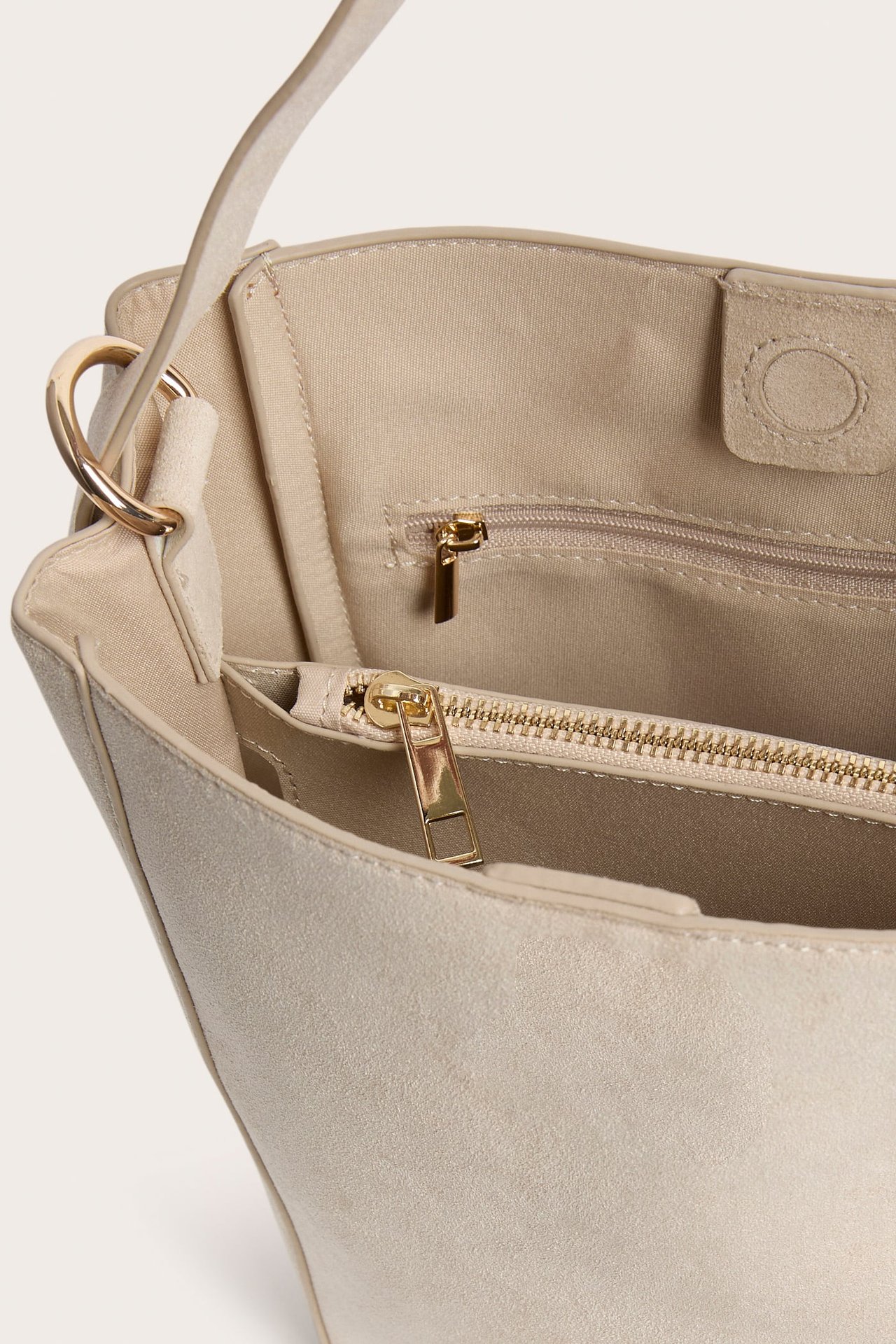Nærbilde av en lys beige bøtteveske i imitert semsket skinn, som viser gullfargede glidelåser og innerfôr.