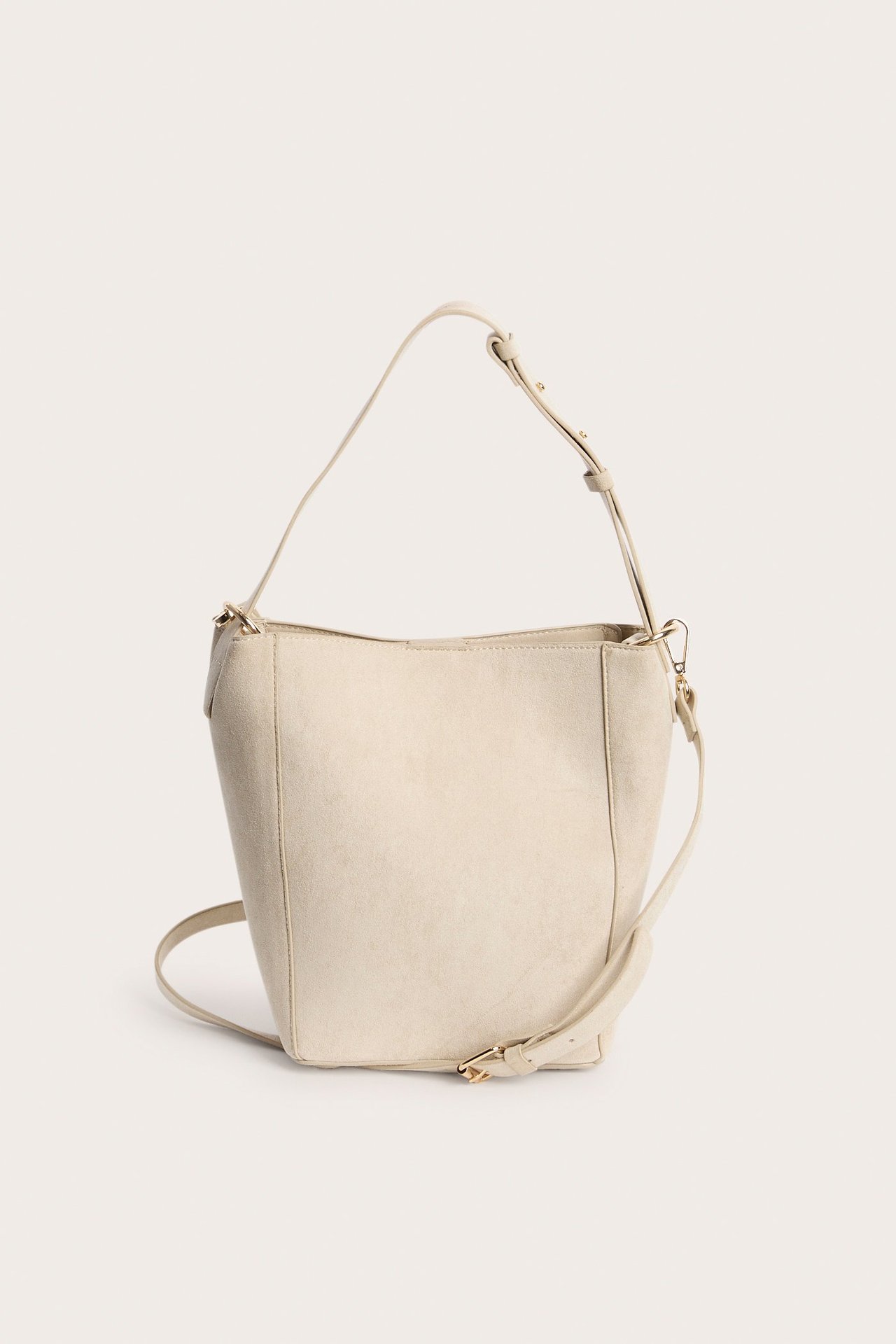 Frontvisning av en lys beige bøtteveske i fuskemocka med justerbar skulderrem og magnetlås.