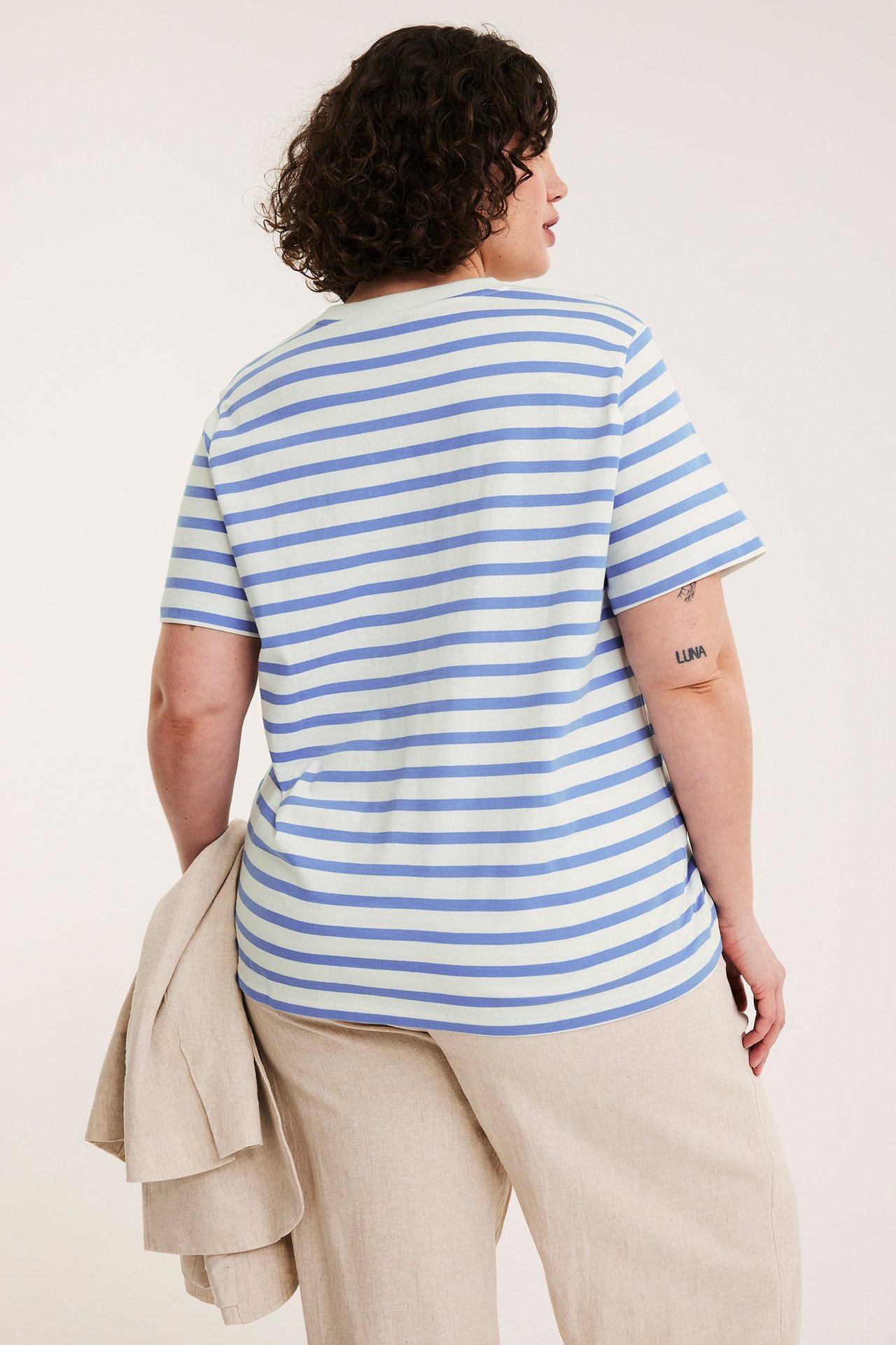 Kvinna sedd bakifrån i ljusblå och vitrandig kortärmad bomulls-t-shirt, beiga byxor, draperad jacka.