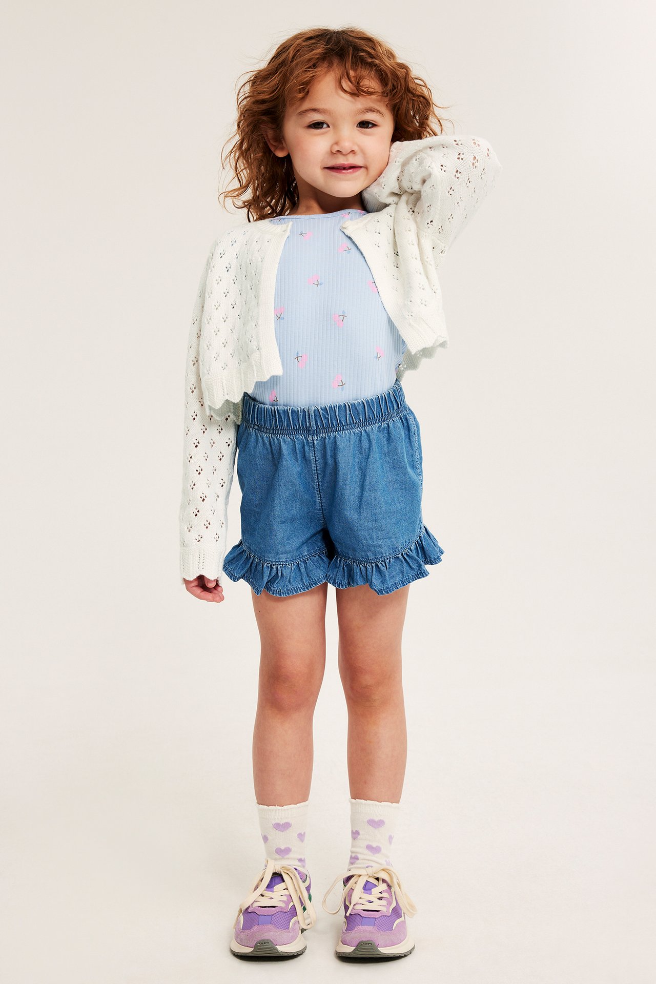Lapsi yllään luonnonvalkoinen reikäneulejakku, vaaleansininen toppi, siniset röyhelöshortsit, sydänsukat ja lila lenkkarit.