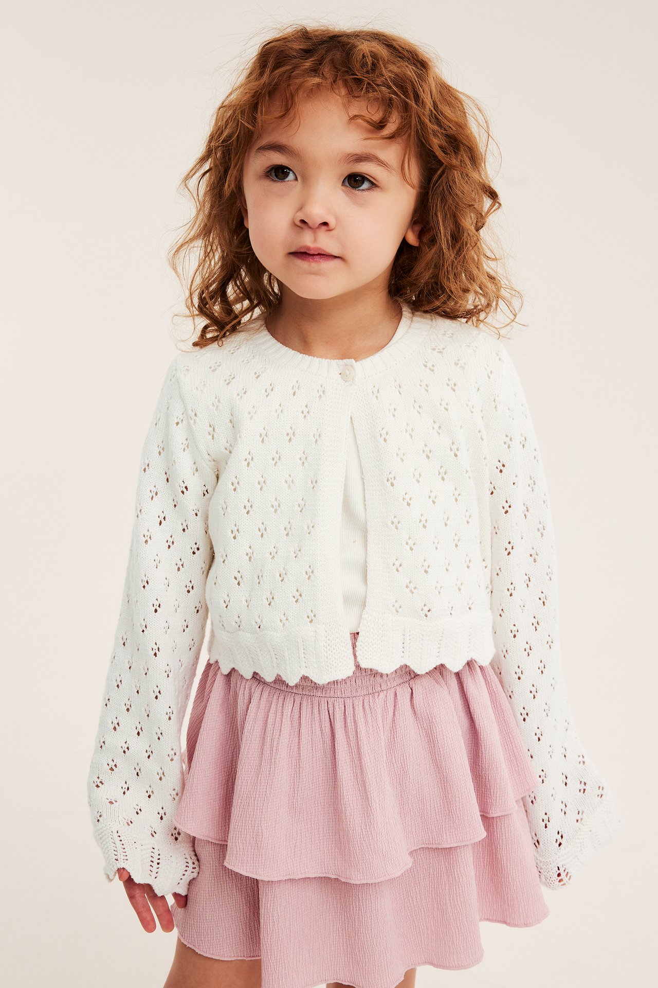 Framifrån: Ett barn bär en offwhite mönsterstickad bomullskofta med vågkant över en ljusrosa volangkjol.