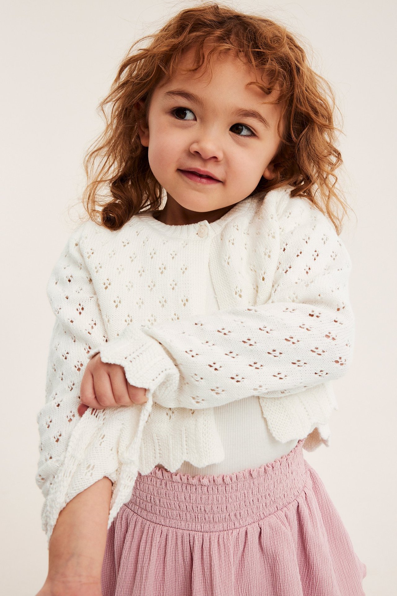 Nærbilde forfra av et barn i en offwhite mønsterstrikket bomullskofta med bølgete kanter over et rosa skjørt.