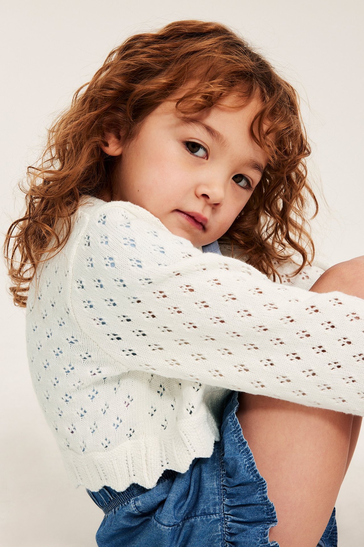 Lapsi yllään luonnonvalkoinen kuviollinen neulottu puuvillatakki aaltoilevalla helmalla, sinisen paidan ja röyhelöfarkkuhameen päällä.