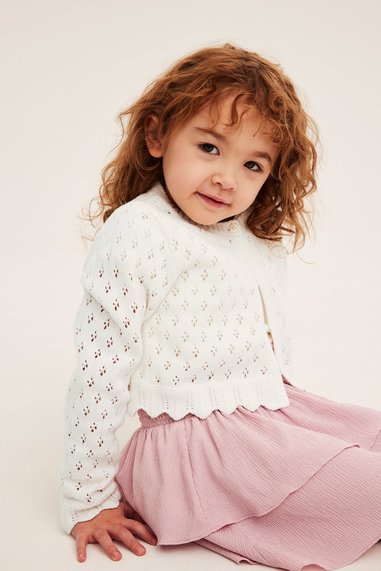 Framsida av ett barn i offwhite mönsterstickad kofta i bomull med vågig nederkant över en rosa kjol.