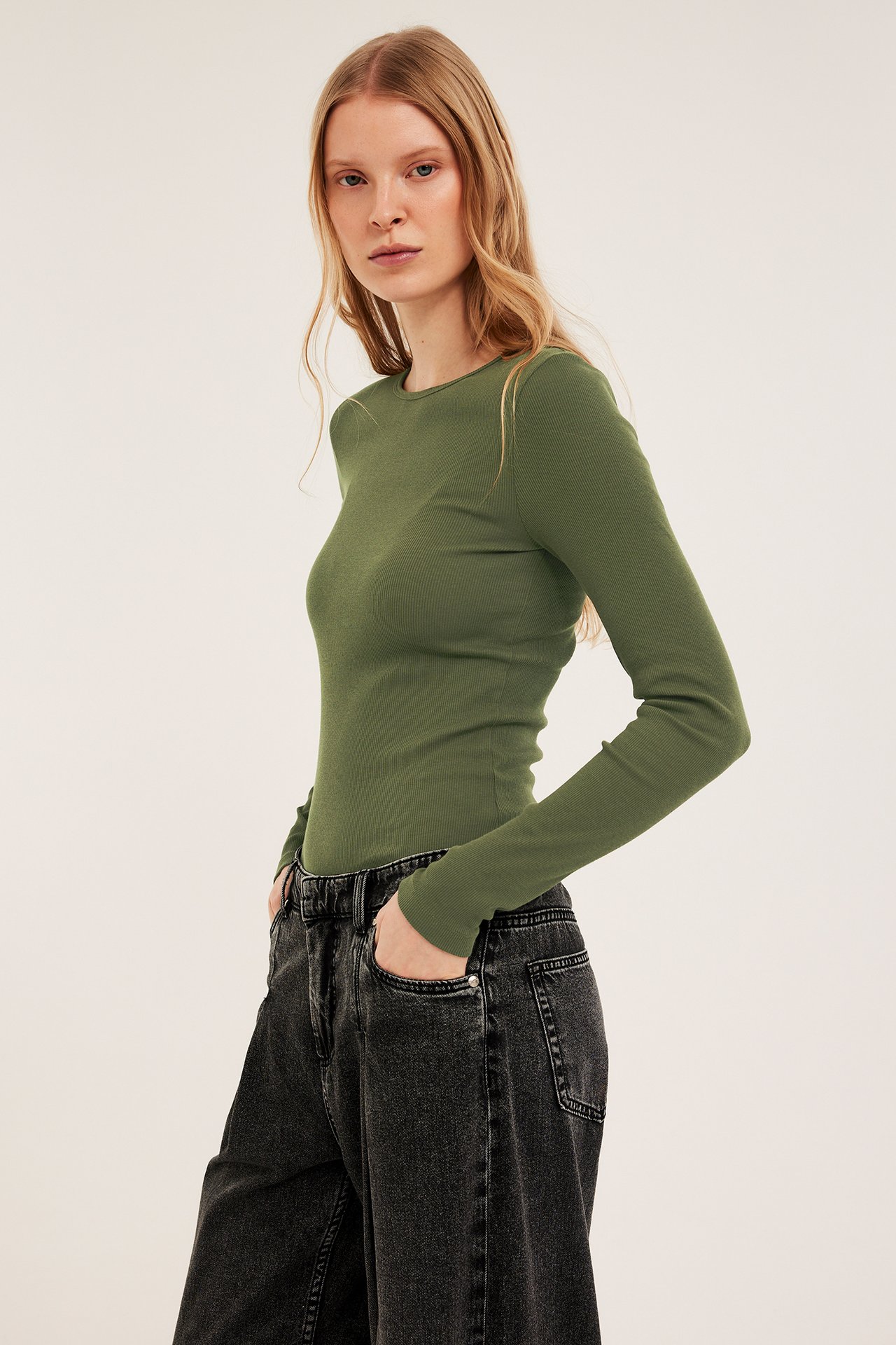Framifrån: Kvinna i en åtsittande khakigrön ribbad långärmad topp och mörkgrå vida jeans.
