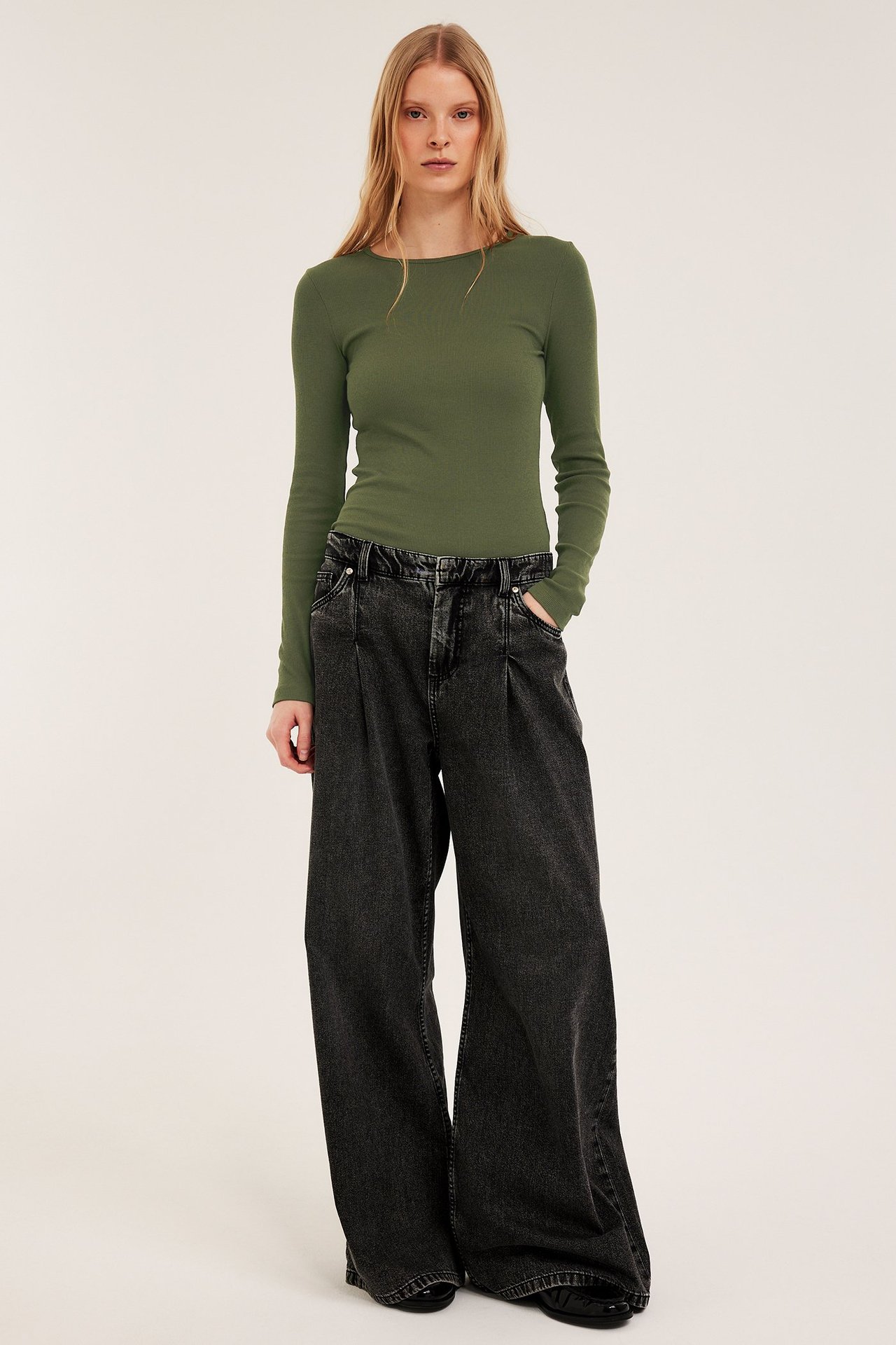 Widok z przodu kobiety w khaki zielonym prążkowanym topie z długim rękawem i ciemnoszarych szerokich dżinsach.