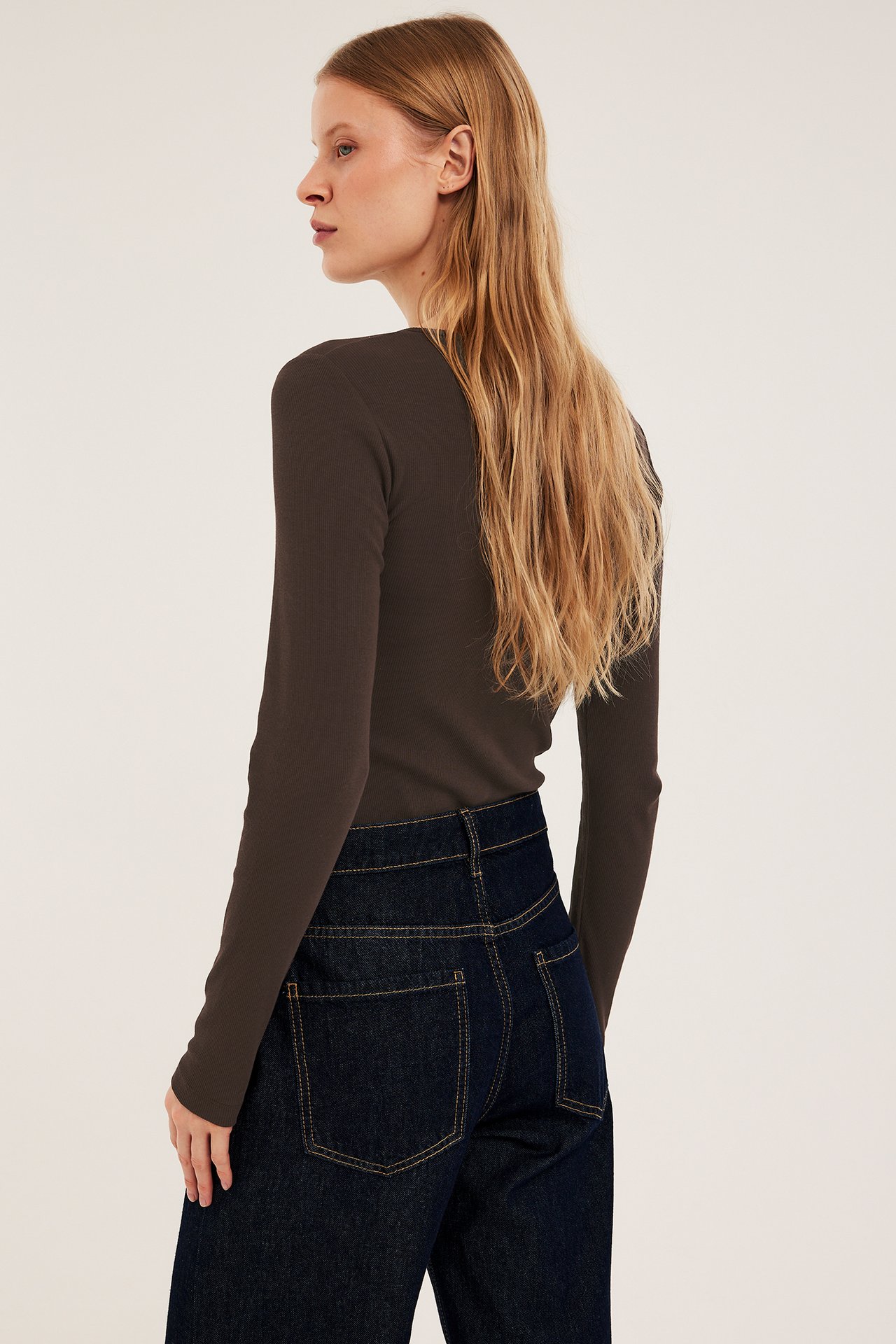 Nainen sivulta takaa, yllään tummanruskea ribattu pitkähihainen toppi ja tummansiniset farkut.