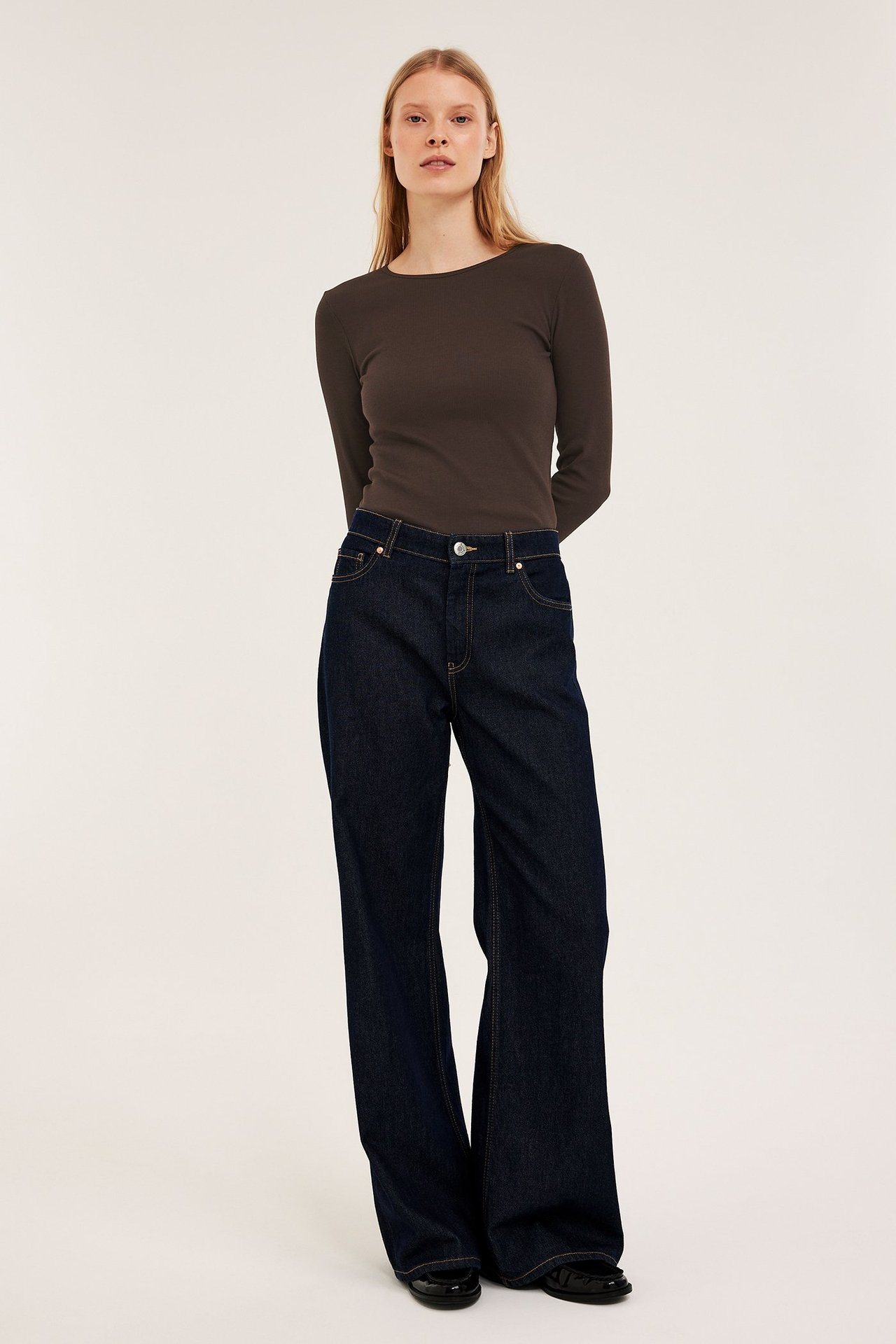 Edestäpäin: nainen pukeutuneena tummanruskeaan ribattuun pitkähihaiseen toppiin ja tummiin leveälahkeisiin farkkuihin.