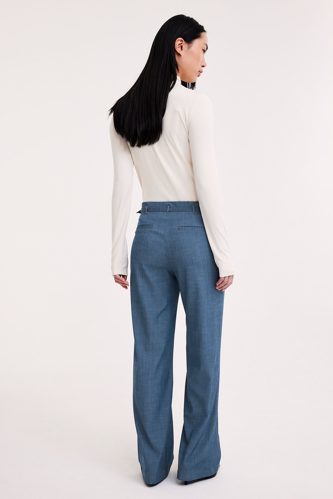 Takaa, nainen yllään vaaleansiniset leveälahkeiset housut säädettävällä vyöllä ja luonnonvalkoinen pitkähihainen paita.
