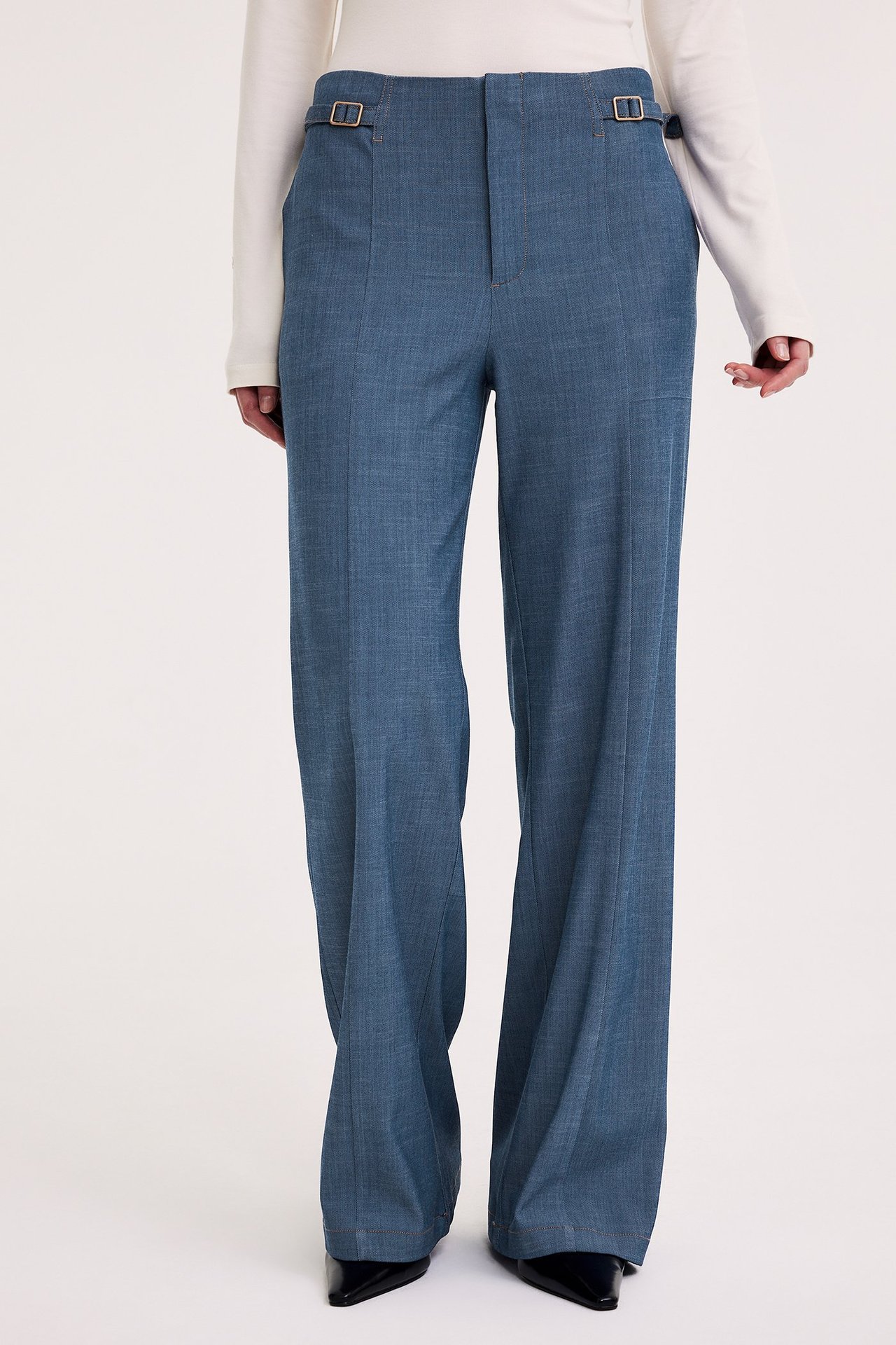 Nainen edestäpäin siniset leveälahkeiset housut vyötärösoljilla, valkoinen toppi ja mustat kärkikengät.