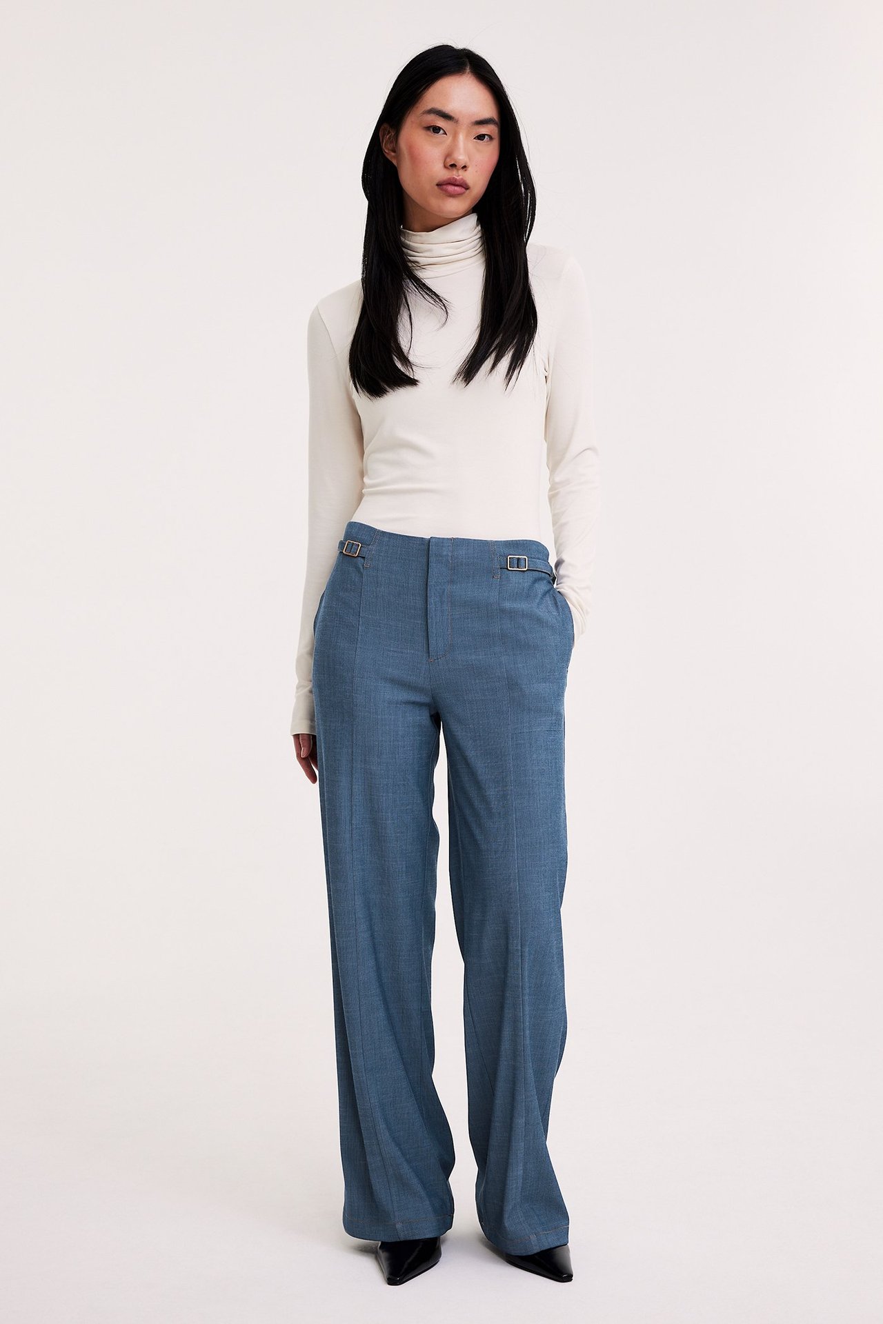 Przód: Kobieta w jasnoniebieskich szerokich spodniach z klamrami, kremowy golf, czarne buty.