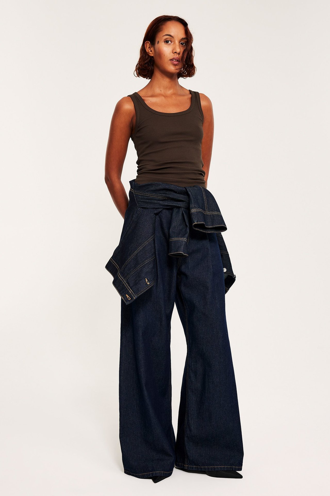 Frontbilde: Kvinne i mørkebrun ribbet singlet, mørkeblå vide jeans og dongerijakke knyttet rundt midjen.