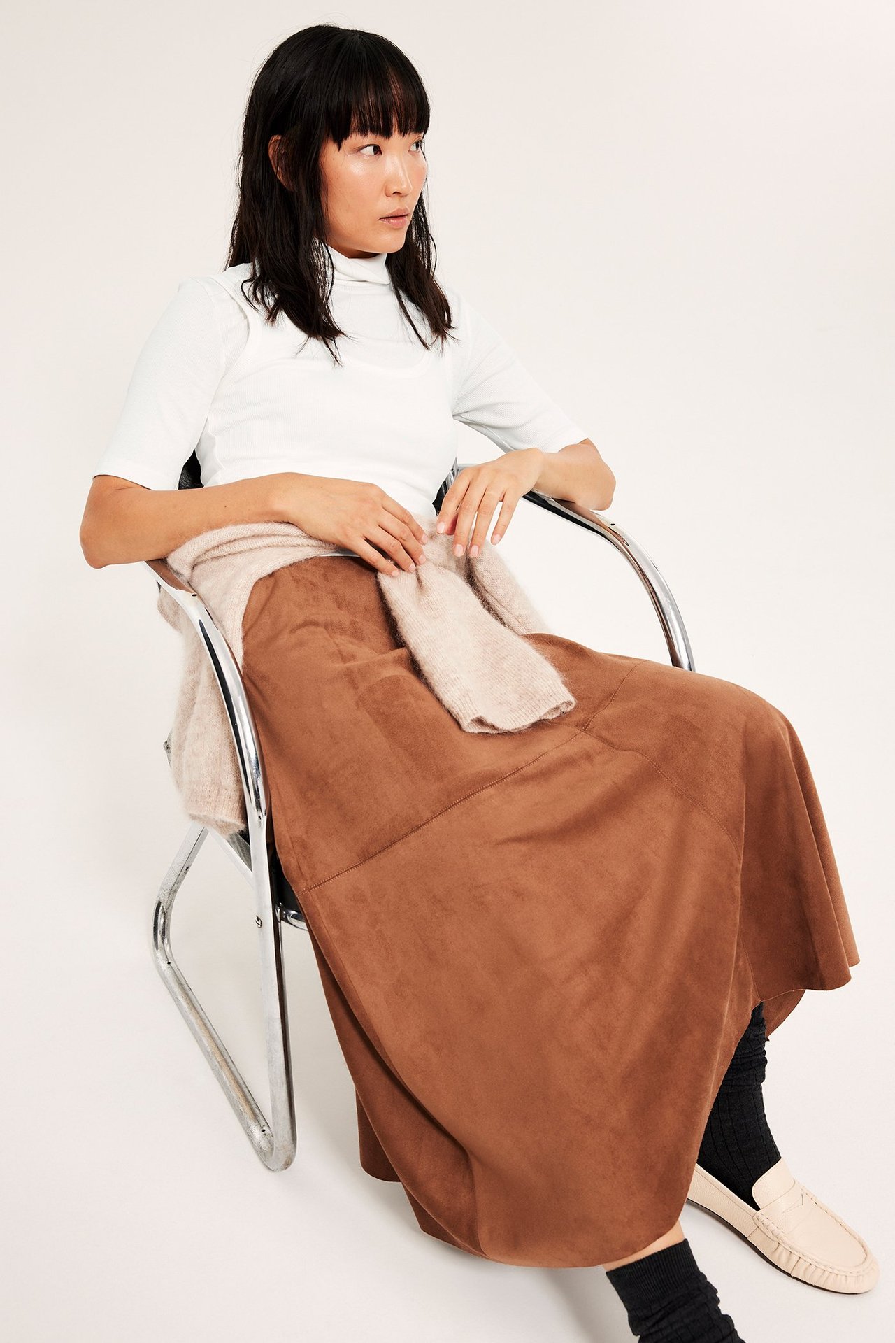 Kvinne i hvitt ribbet topp med høy hals, sitter, med beige genser og brun skjørt.