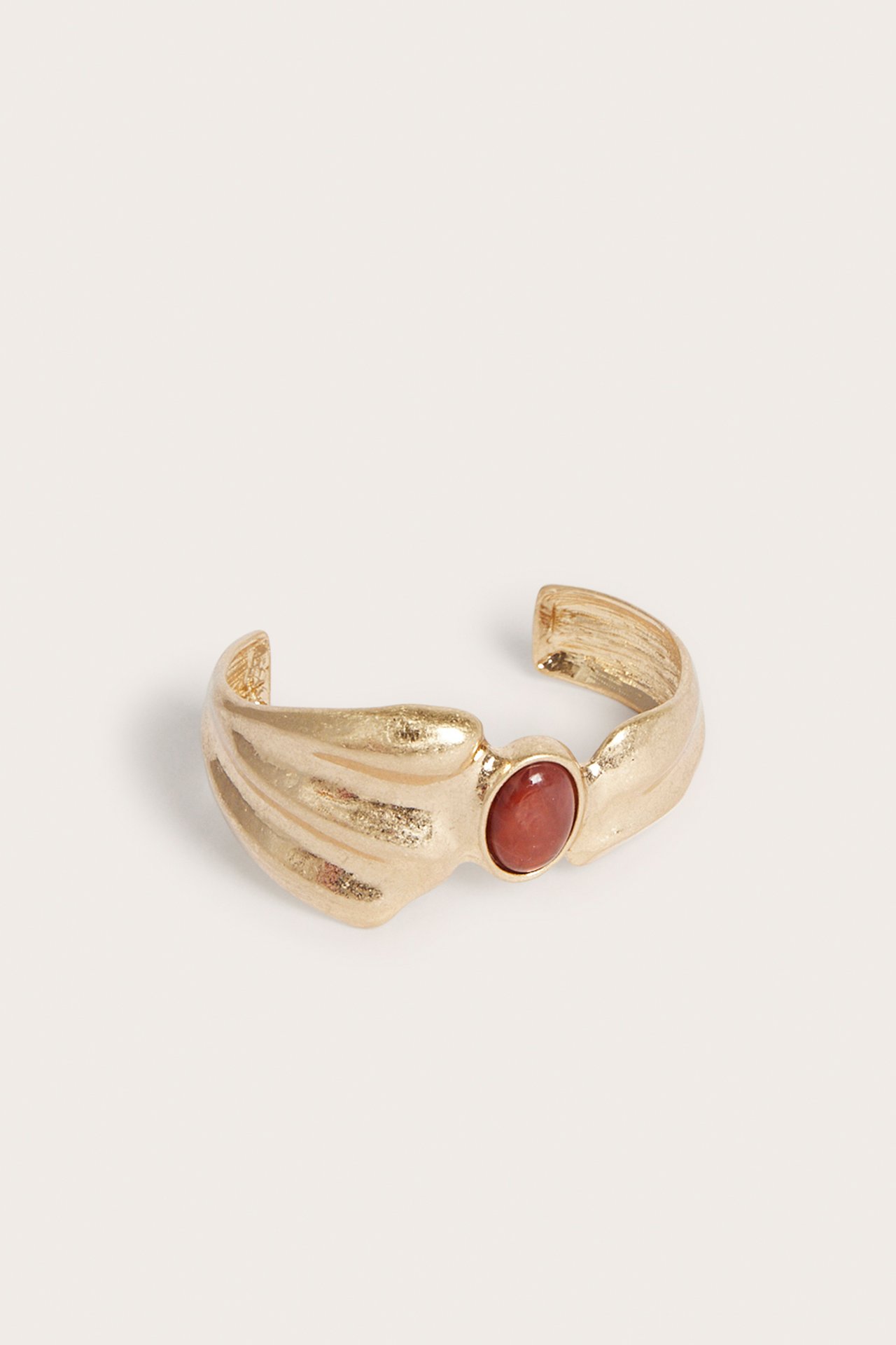 Närbild framifrån: Ett guldfärgat texturerat stelt armband med en röd plaststen.