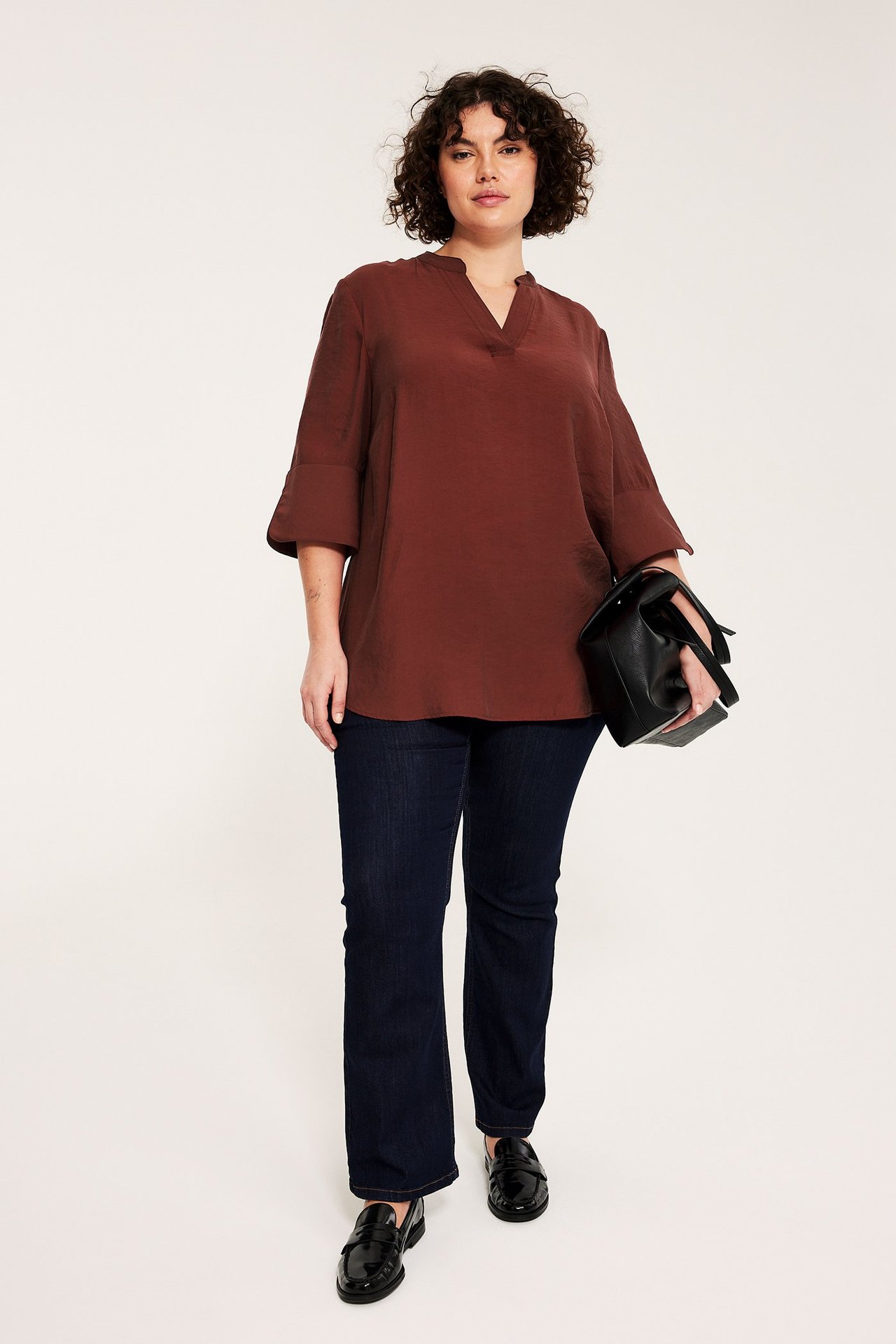 Kobieta w czerwonej bluzce V-neck, granatowych dżinsach, czarnych mokasynach, z czarną torebką. Widok z przodu.