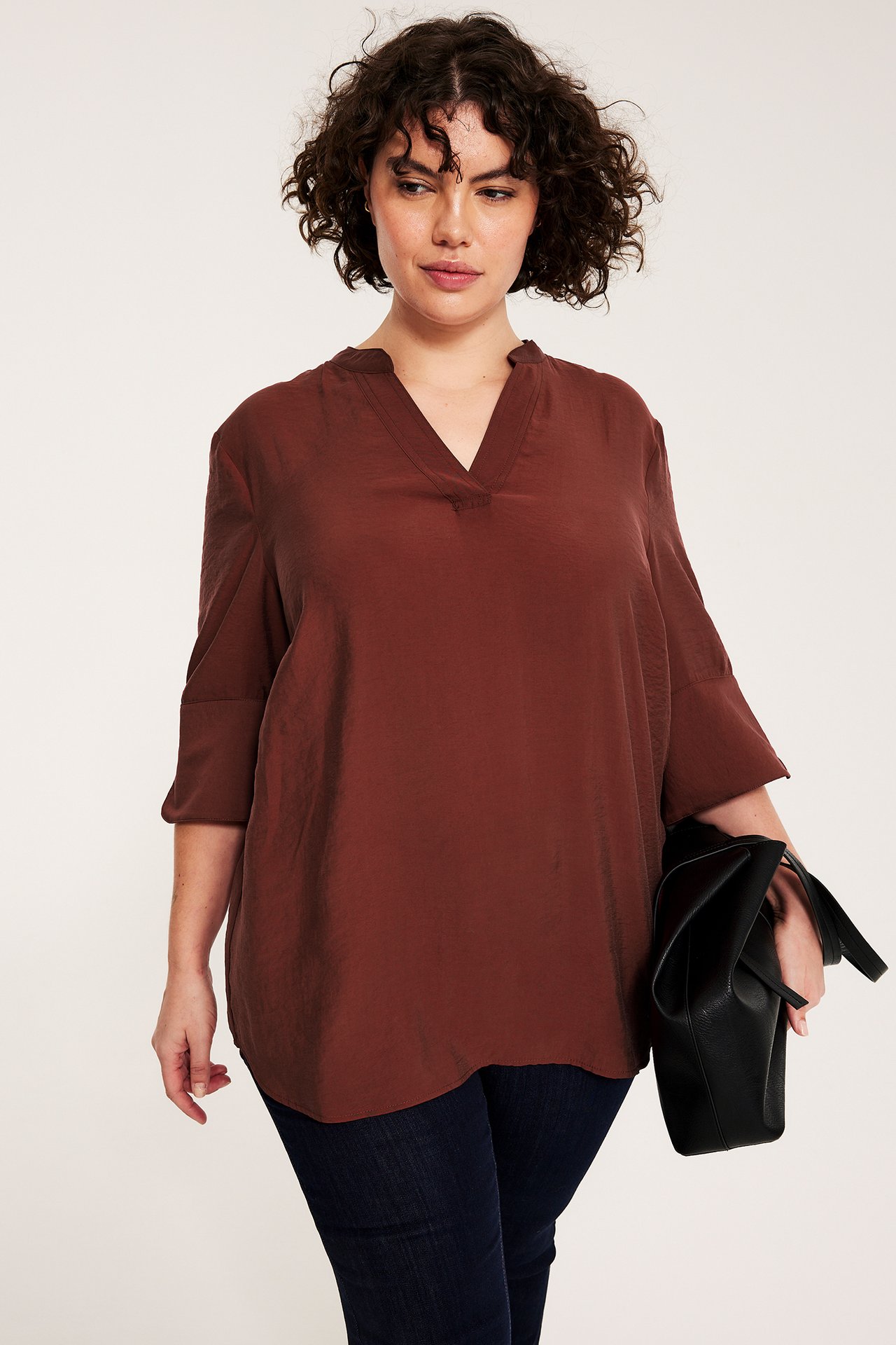 Framifrån: Kvinna i lös röd sidenskimrande V-ringad blus, mörka jeans, hållande en svart handväska.