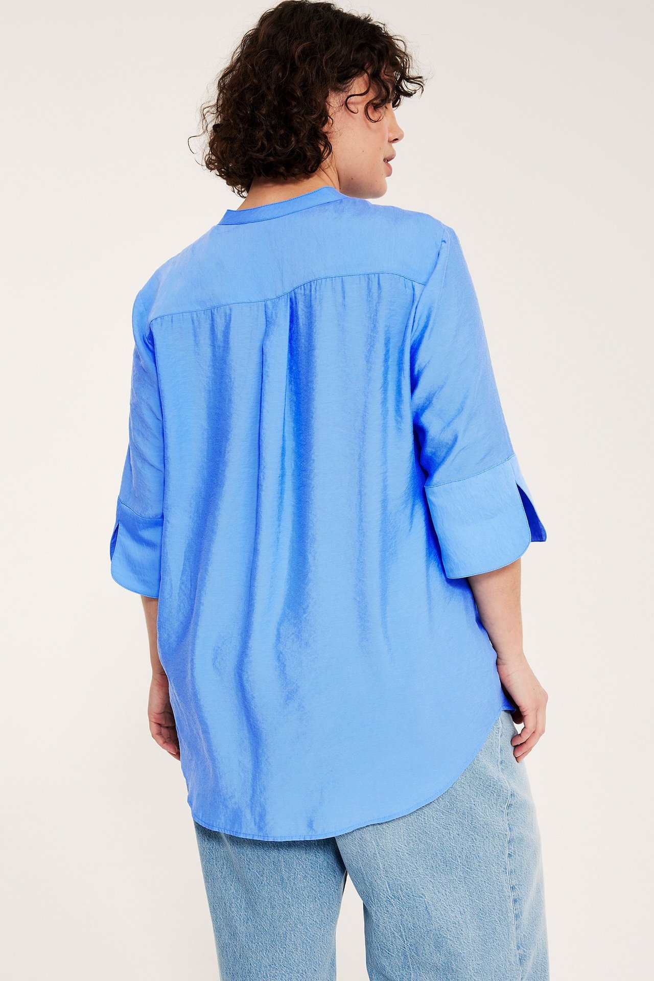 Kobieta, widok z tyłu: niebieska jedwabna bluzka z podwiniętymi rękawami 3/4 i jasnoniebieskie dżinsy.