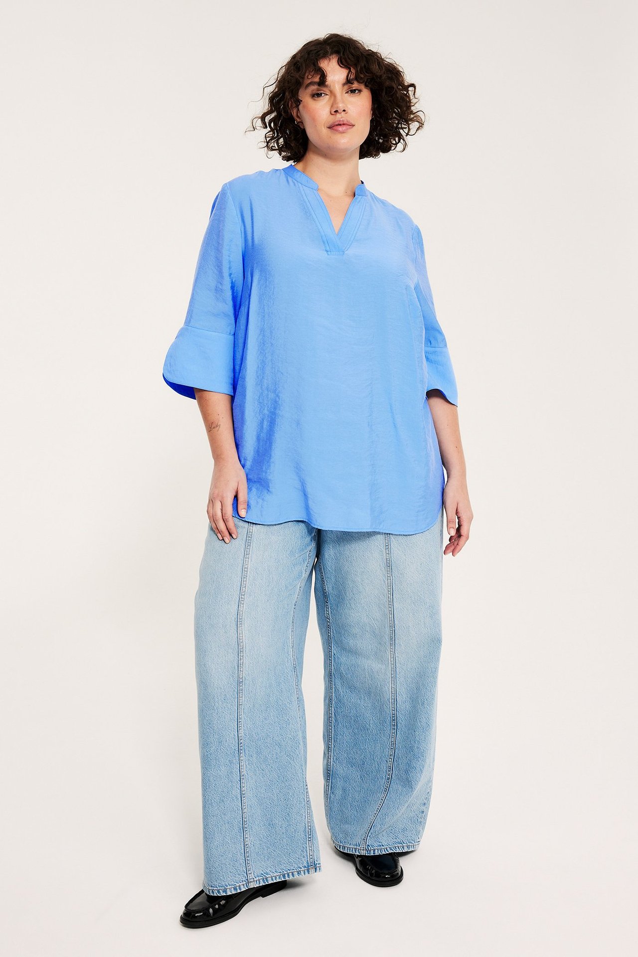 Widok z przodu kobiety: niebieska bluzka V-neck, jasnoniebieskie szerokie dżinsy, czarne mokasyny.