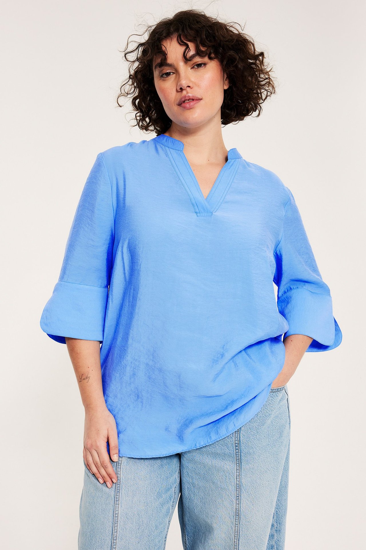 Kobieta z przodu: niebieska bluzka z dekoltem w serek, rękawami 3/4 i jasne dżinsy.