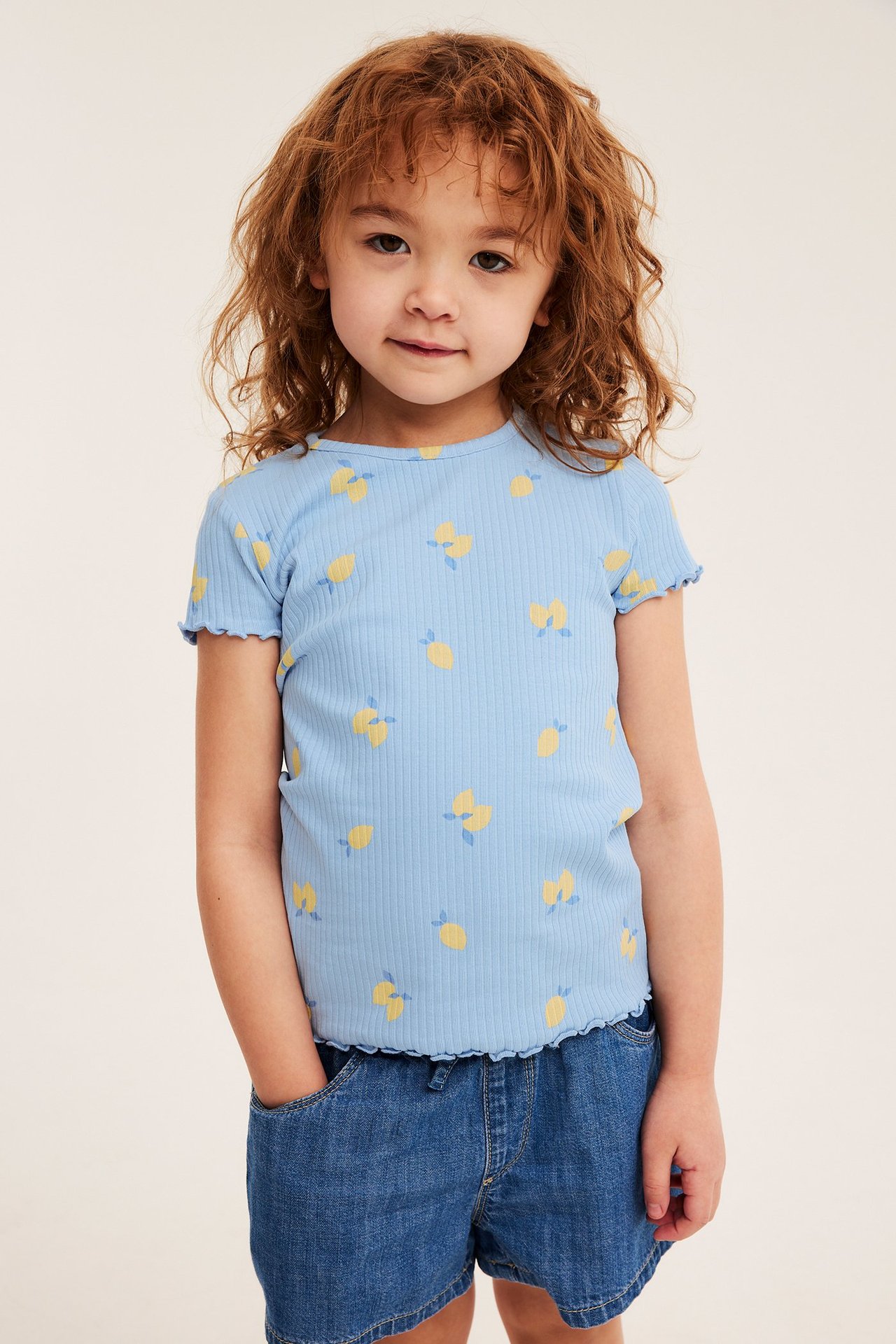 Frontvy på ett barn i ljusblå ribbad kortärmad topp med citronmönster och blå denimshorts.