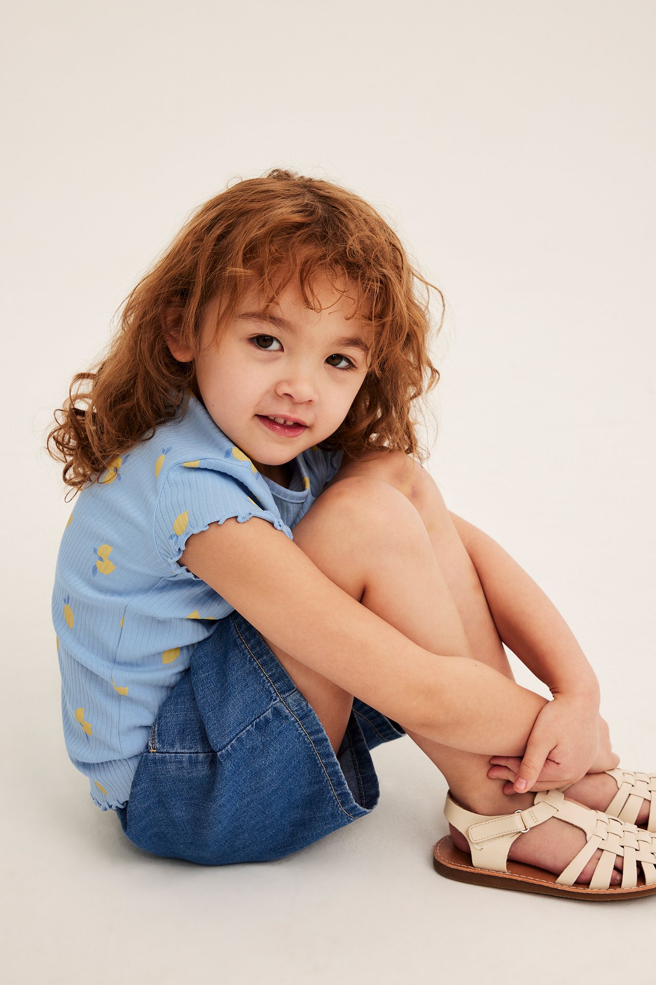 Frontvy av ett barn i en ljusblå ribbad kortärmad topp med gula citroner, en jeanskjol och beiga sandaler.