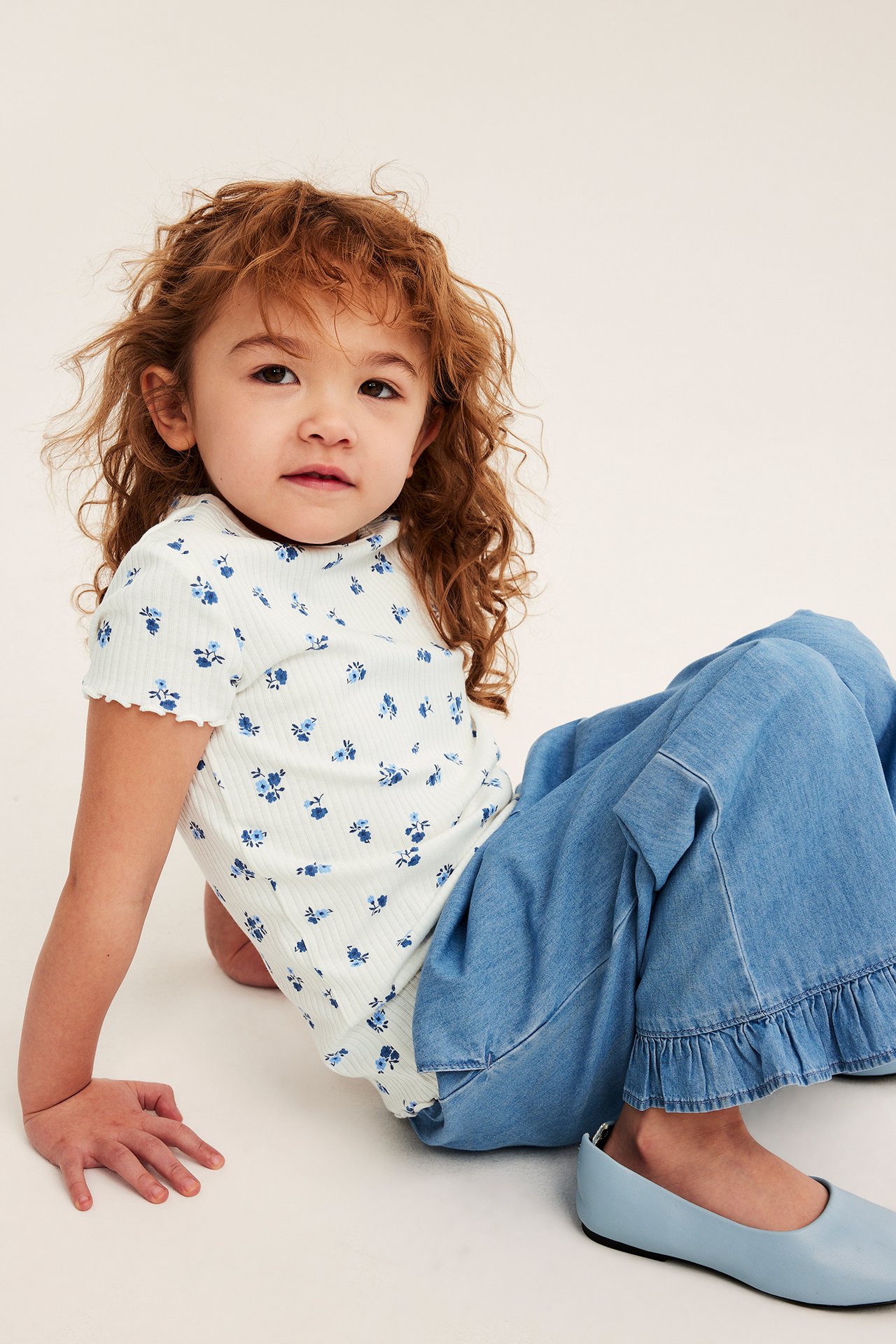 Framifrån: ett barn i offwhite ribbad kortärmad blommig topp, blå vida jeans och ljusblå ballerinaskor.