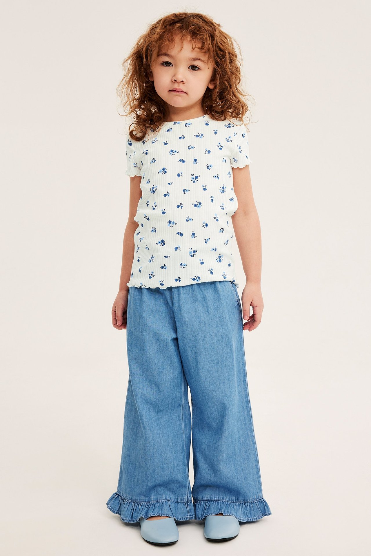Widok z przodu: Dziecko w prążkowanym, kwiecistym topie ecru, niebieskich szerokich spodniach i jasnoniebieskich butach.