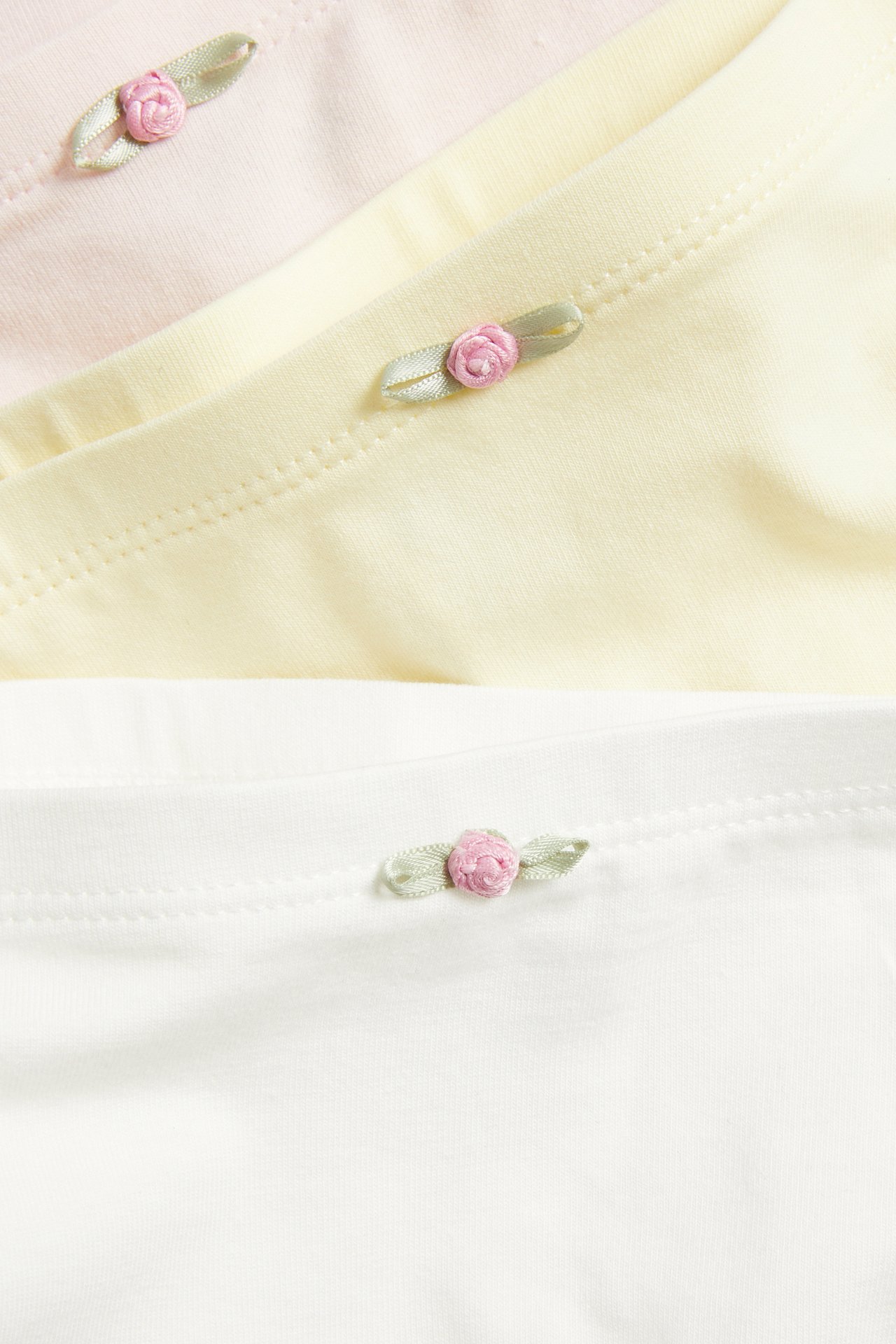 Närbild på tre barnboxertrosor i offwhite, ljusgul och ljusrosa med en liten rosettapplikation.