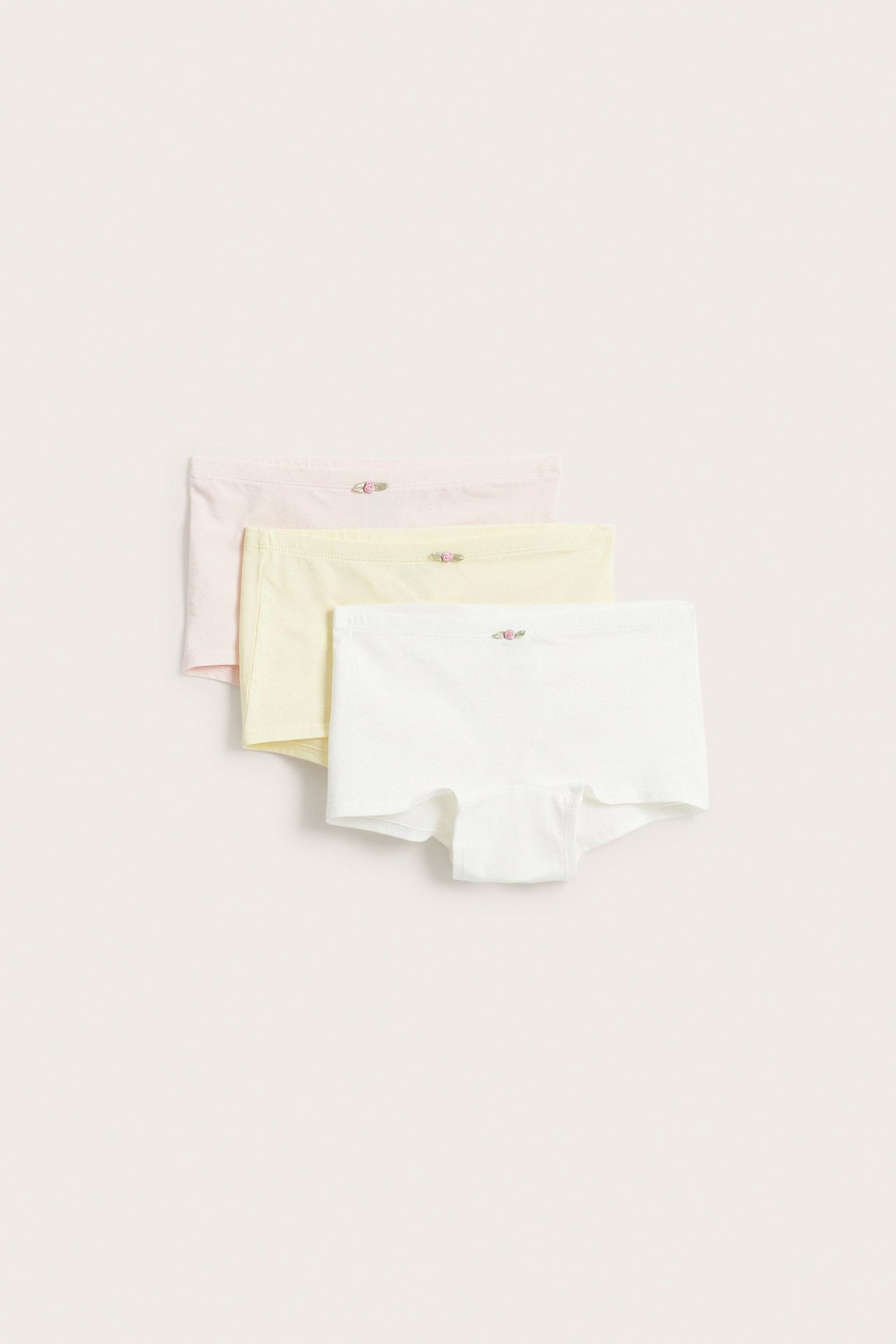 Edestä, kolme lasten puuvillabokseria: luonnonvalkoinen, vaaleankeltainen ja vaaleanpunainen, kussakin ruusuapplikaatio.